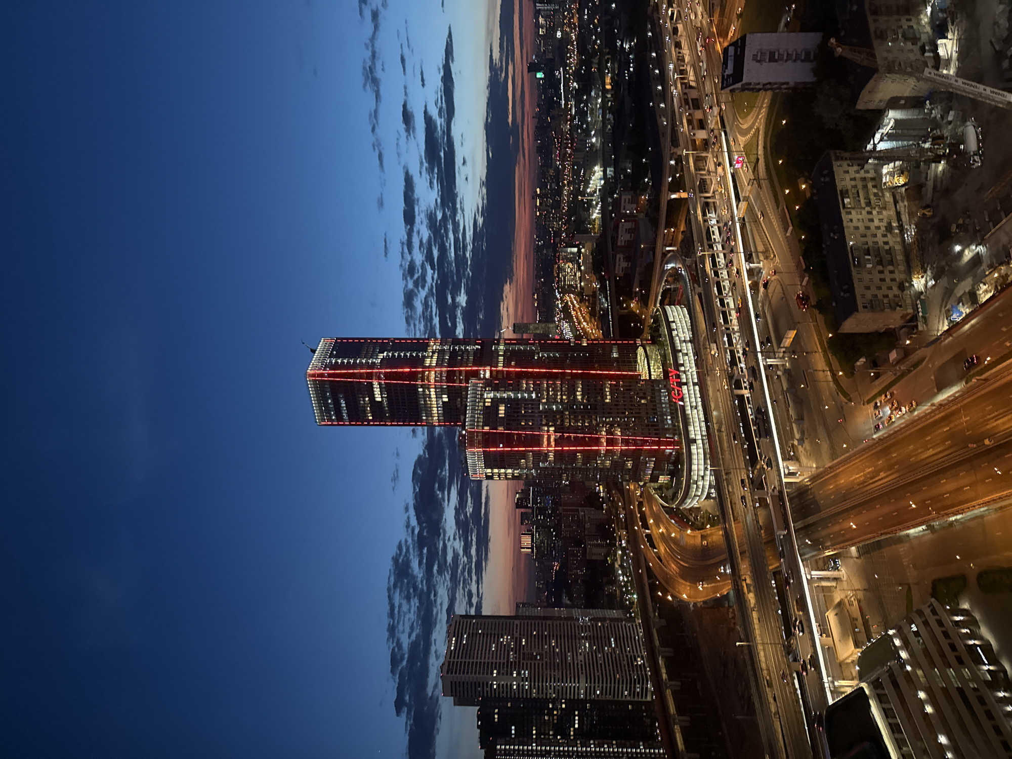 The image captures an aerial or high-angle view of a city, identified as Khimki, Russia, at dusk or early night. The sky transitions from a deep blue overhead to a lighter, warm-hued horizon, with distinct cloud formations.
In the center-left, two prominent modern skyscrapers are a focal point, characterized by striking vertical red LED lights running along their facades. The rightmost of these two towers displays the text "iCITY" near its top. Surrounding these high-rises are numerous other buildings, both residential and commercial, with many windows illuminated.
Below and to the right, a complex and extensive network of highways and roads is visible, brightly lit by streetlights. Numerous cars are present, their headlights and taillights appearing as trails or individual points of light, indicating active traffic flow. The city lights extend far into the distance, illuminating the urban sprawl. In the foreground on the right, what appears to be a construction site with cranes can be observed. The scene conveys the vibrant, illuminated atmosphere of a large city after sunset.