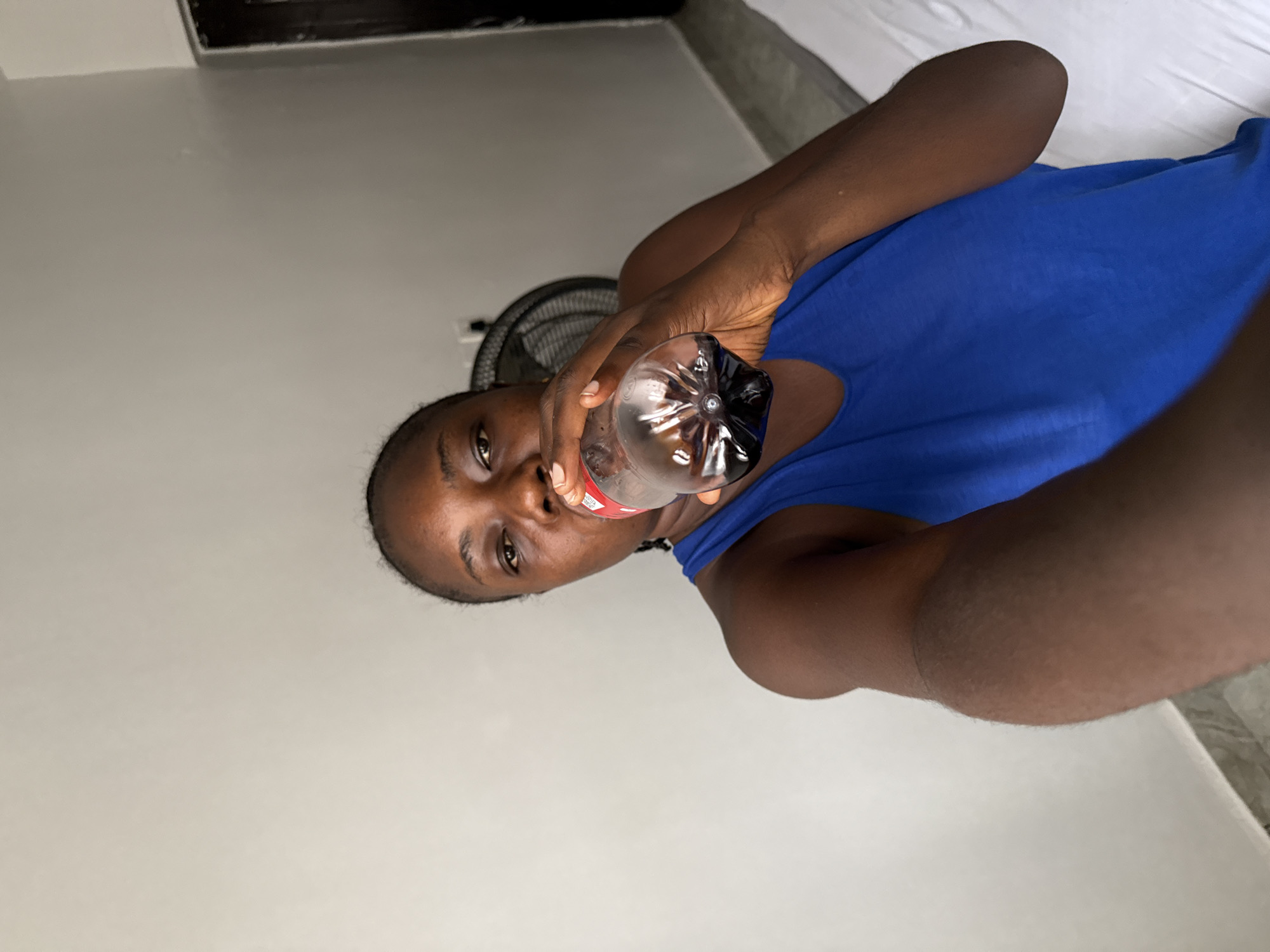 A person wearing a blue tank top is drinking from a plastic Coca-Cola bottle. A black fan is visible in the background. The location is in an indoor room in Iddo Tudunwada, Nigeria. The surroundings appear to be domestic.