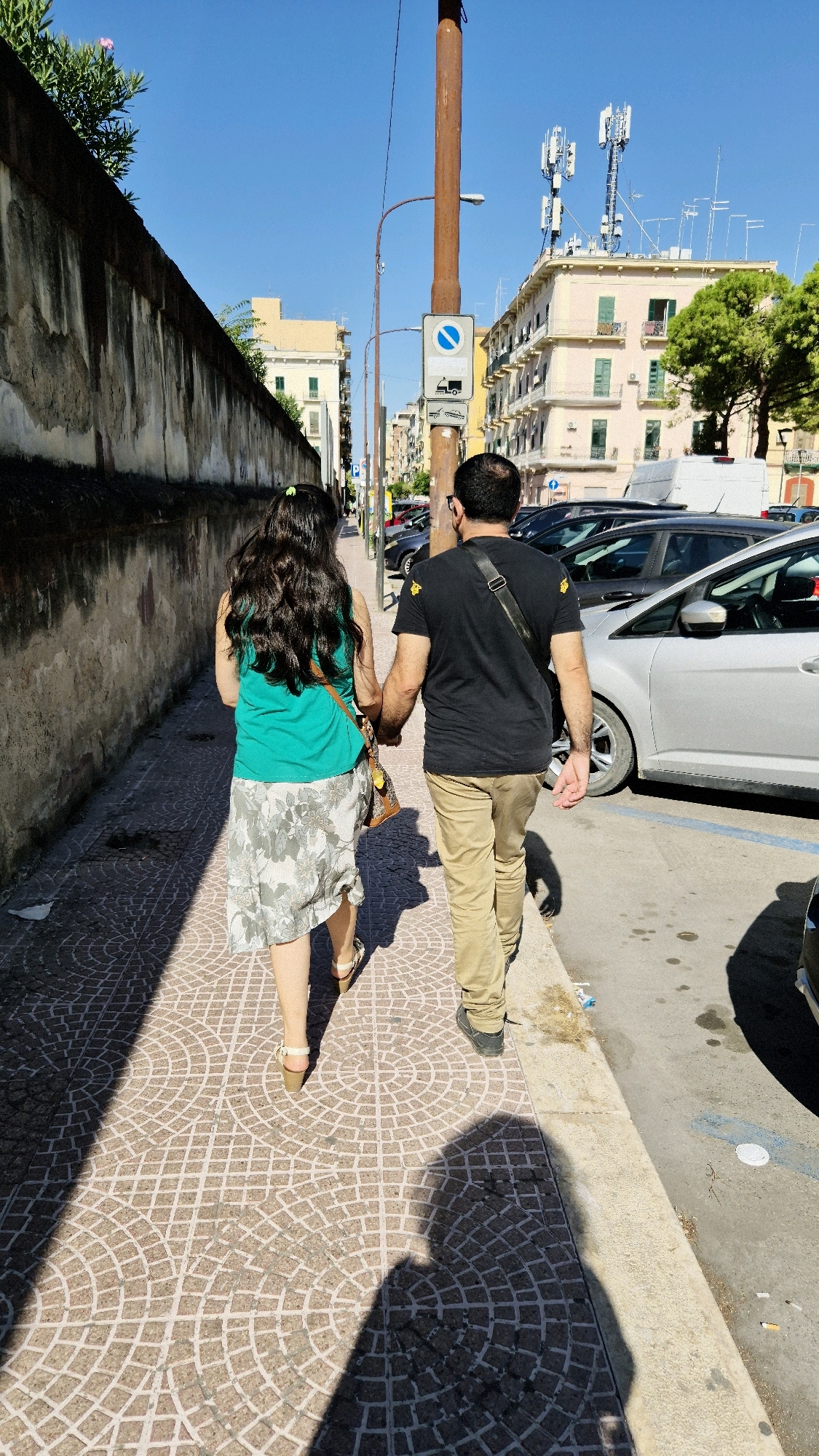 A man and a woman are walking hand-in-hand on a sun-drenched sidewalk in Taranto, Italy. The woman, seen from behind, has long, dark, wavy hair and wears a green tank top and a floral print skirt. She has a brown bag slung over her shoulder. The man, also viewed from behind, wears a black t-shirt with yellow accents and khaki pants. He has a dark strap across his chest, likely from a bag. They are walking away from the camera, casting long shadows on the patterned cobblestone sidewalk.
To their left is a tall, weathered stone wall. To their right, parked cars line the street. In the background, a light pink building with green shutters stands out, topped with numerous telecommunication antennas. The sky is a clear, bright blue. The overall scene depicts a casual stroll on a warm day in an urban environment.