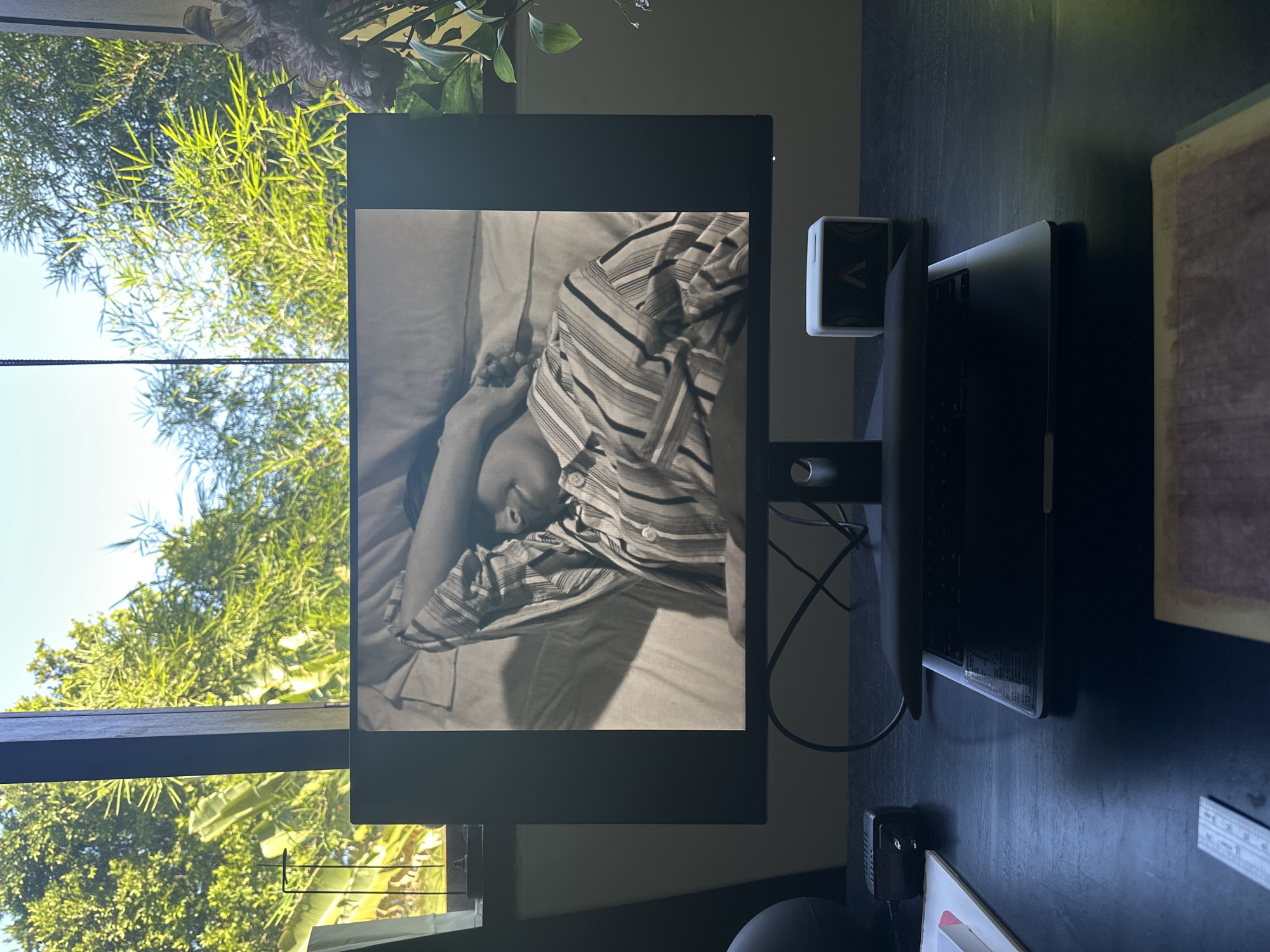 A monitor displaying a black and white image of a person in a striped shirt lies on a bed. The monitor sits on a dark-colored desk with other items, including a small white cube and a keyboard. Outside a window, lush green vegetation is visible. The location is North Kuta, Indonesia.