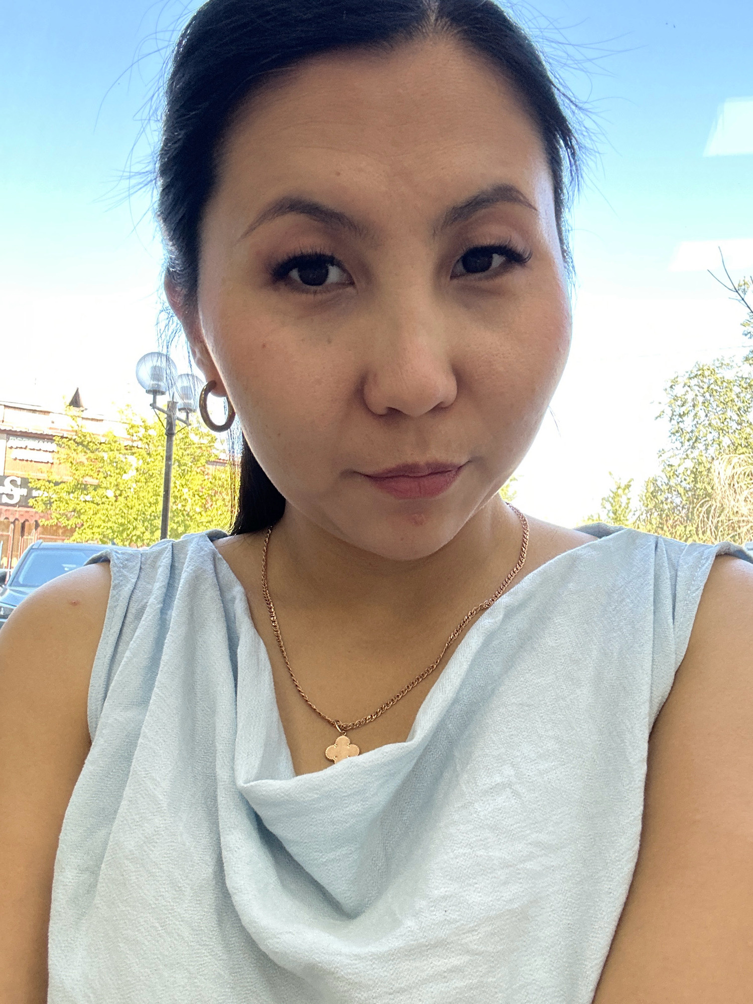 This is a medium close-up selfie of a woman with dark hair, wearing a light blue sleeveless top and a gold chain necklace with a clover-shaped pendant. She has makeup on, including mascara and blush. She is wearing a gold hoop earring in her left ear. The background shows a bright blue sky, some greenery, and a lamppost with buildings in the distance. The overall setting appears to be outdoors in a city.