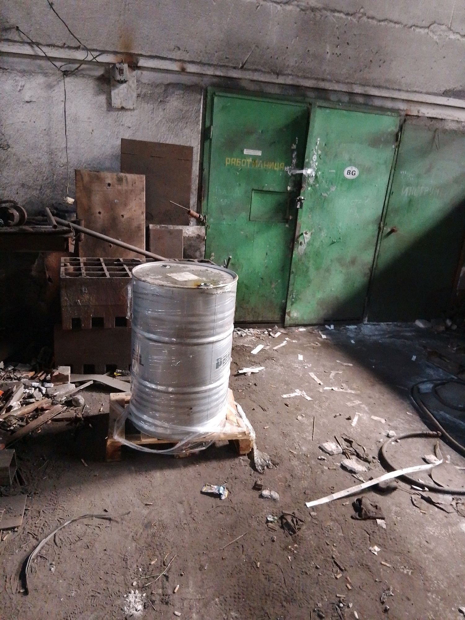 The image depicts a cluttered industrial or storage space. In the foreground, a large metal barrel, wrapped in plastic, sits on a wooden pallet. The floor is dirty and strewn with debris, including wood scraps, metal pieces, and what appears to be cigarette butts. To the left of the barrel, there are stacks of industrial materials, possibly metal or concrete blocks, and some tools. In the background, two large green metal doors are visible, one of which has a circular sticker with the letters "BG" inside. Text is visible on the left door, reading "РАБОТЯНИЦА" (rabotyanitsa), which translates to "workwoman" or "workroom." The walls are rough and textured, and electrical wires are visible overhead. The lighting suggests an indoor, possibly dimly lit environment.