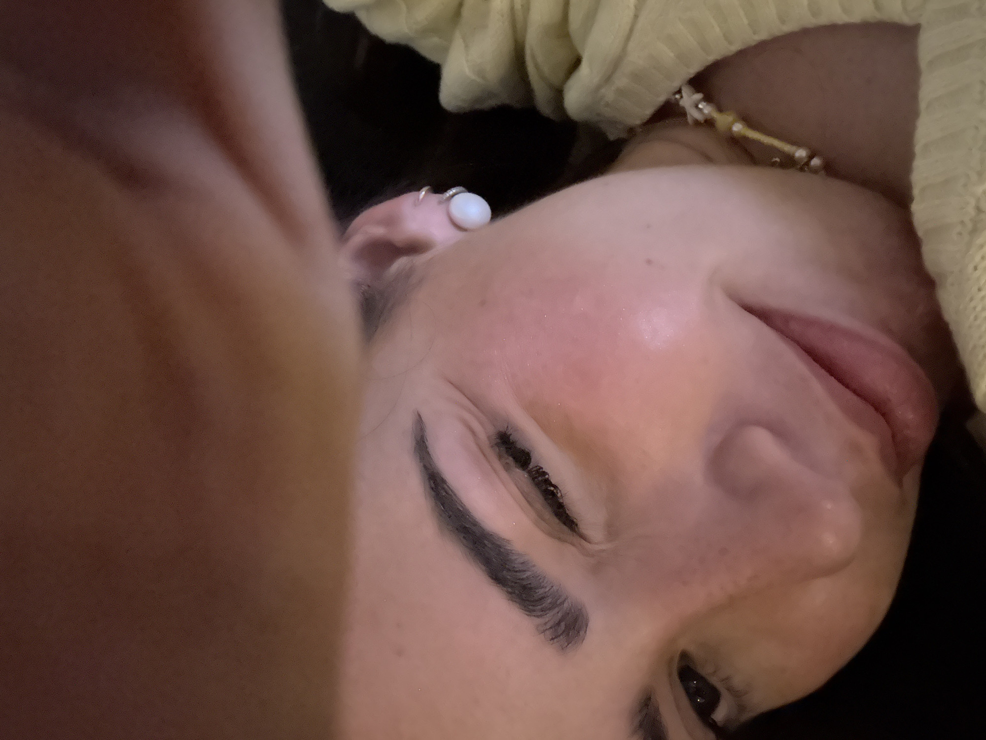 A close-up, tilted view shows a person's face. The person has fair skin, dark eyebrows, and closed eyes. A white earring is visible on the ear. Part of a yellow, knit garment is seen in the upper right corner. The setting or location details for San Bernardino Tlaxcalancingo, Mexico, are not discernable from the image provided.