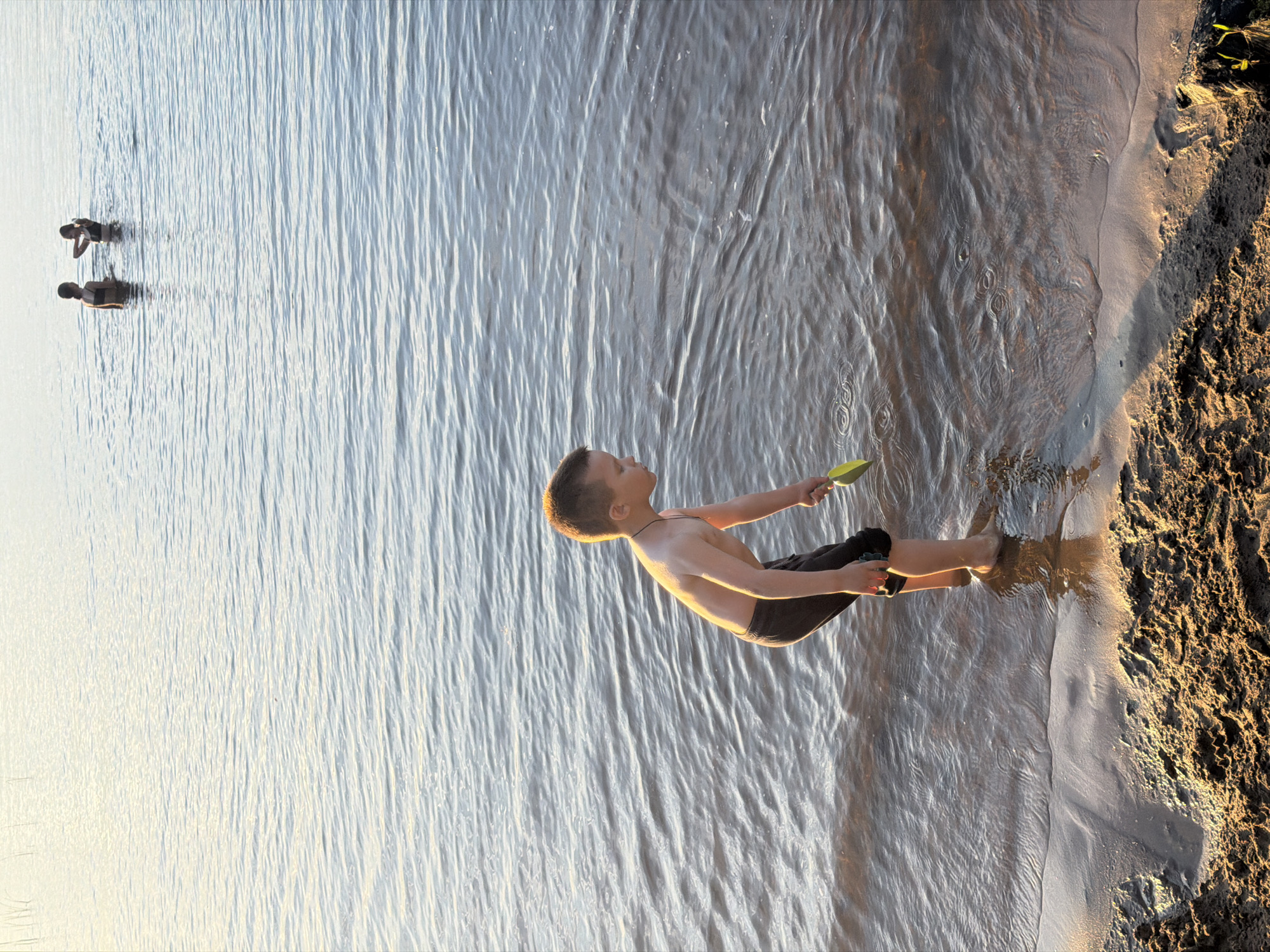 The image depicts a serene natural scene, likely a beach or shoreline, with calm water reflecting the soft, diffused light of what appears to be late afternoon or early morning. The water is a light, hazy blue-gray, with gentle ripples disturbing its surface. In the distance, three figures are visible in the water, two of whom are partially submerged and facing towards the foreground. In the foreground, a young boy stands at the edge of the water, his body angled to the right. He is shirtless and wearing dark shorts, his wet feet submerged in the shallow water. He holds a bright yellow shovel in his right hand, which is positioned as if he is about to scoop something from the water or sand. The shoreline is a mix of wet sand and darker, coarser sand or mud, with some texture and undulations. The overall impression is one of peaceful recreation in a natural environment. This scene could potentially be located on the shores of the Kama River in Chaikovsky, Russia, known for its natural beauty and recreational opportunities.