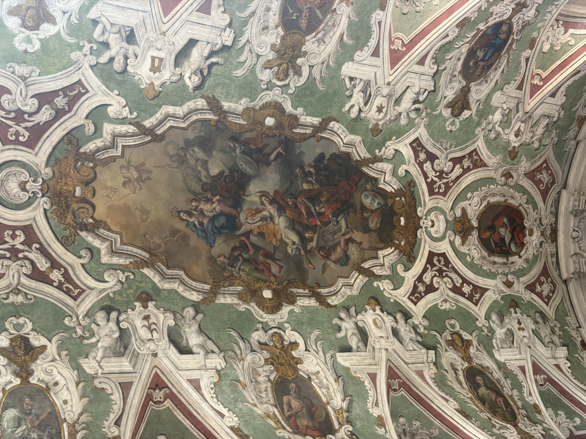 This image depicts a detailed fresco on the ceiling of a room, likely a historical building in Lisbon, Portugal. The fresco is an elaborate baroque-style painting featuring a central oval composition surrounded by intricate stucco work and smaller oval paintings.
The central painting is a religious scene, possibly depicting the Assumption of Mary or a similar heavenly ascension. It shows a group of figures, including women and children, ascending towards a central female figure crowned with a halo and holding a child, all surrounded by cherubs and angels. The figures are dressed in flowing robes, and the overall color palette is rich with blues, golds, reds, and flesh tones.
The surrounding stucco work is elaborate, featuring swirling acanthus leaves, floral motifs, and putti (cherubic figures) in various poses, some appearing to be supporting or interacting with the painted elements. The ceiling is painted in sections, with a dominant green background in the wider areas, bordered by ornate red and white decorative elements.
Scattered throughout the ceiling are several smaller oval paintings, each containing a single figure or a small group. These figures appear to be saints or biblical characters, depicted in traditional religious attire. For instance, one oval painting shows a figure in red robes holding a book, and another displays a scene with figures in drapery.
The overall impression is one of grandeur and opulent artistic decoration, characteristic of religious or noble architecture from the baroque period. Without specific identifying markers within the image itself, pinpointing the exact location in Lisbon is not possible based solely on the visual information provided. However, the style strongly suggests a significant historical or ecclesiastical site.