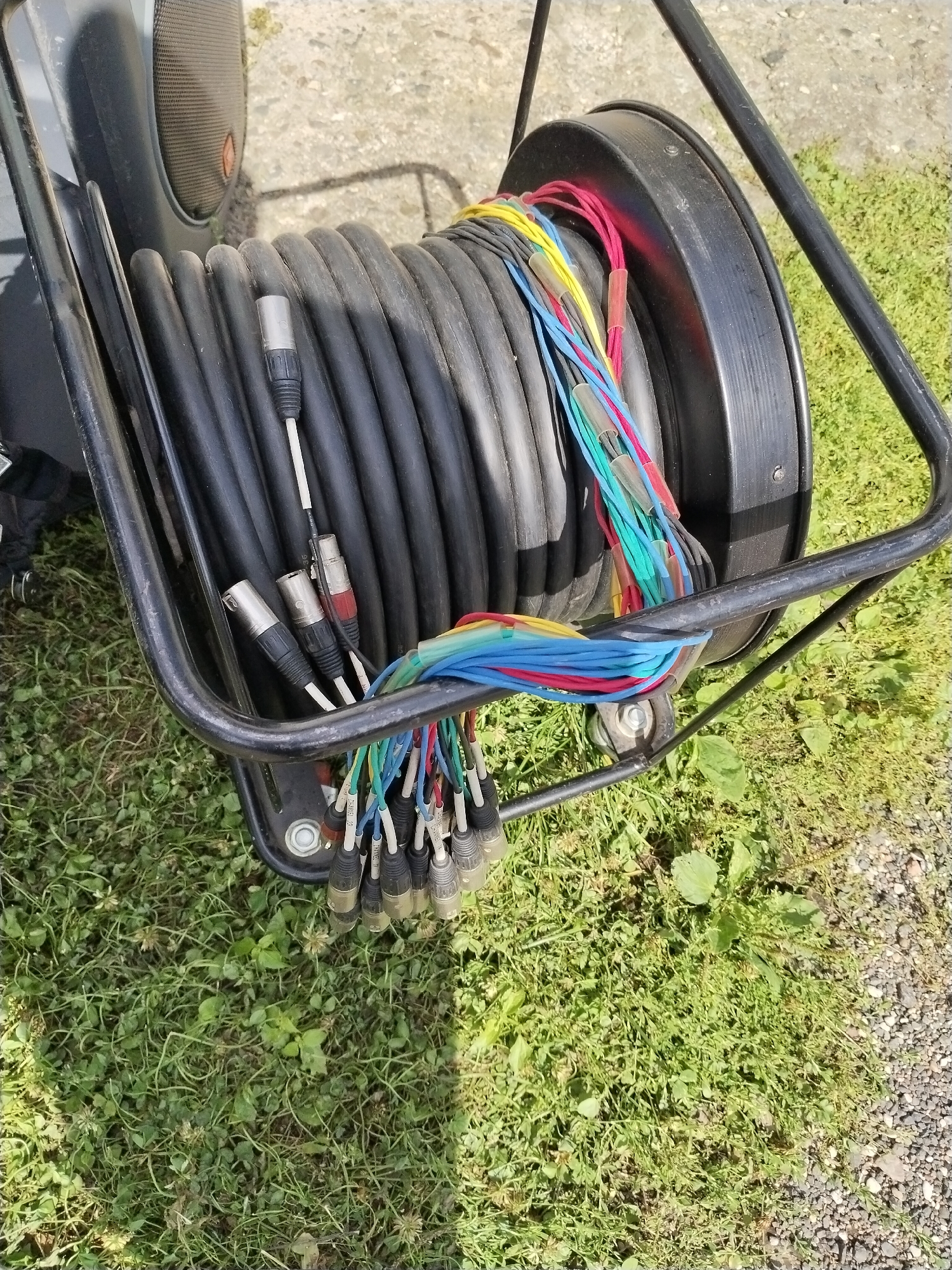 The image shows a coiled-up audio cable on a reel, which is part of a black metal cart. The reel is mostly black, with some colored wires protruding from it. The cart is outdoors, on a grassy area with some gravel. The overall scene appears to be in a city park or a similar outdoor public space. There are no people or animals visible in the image. The image does not provide any information that would help determine the specific location in Hrodna, Belarus.