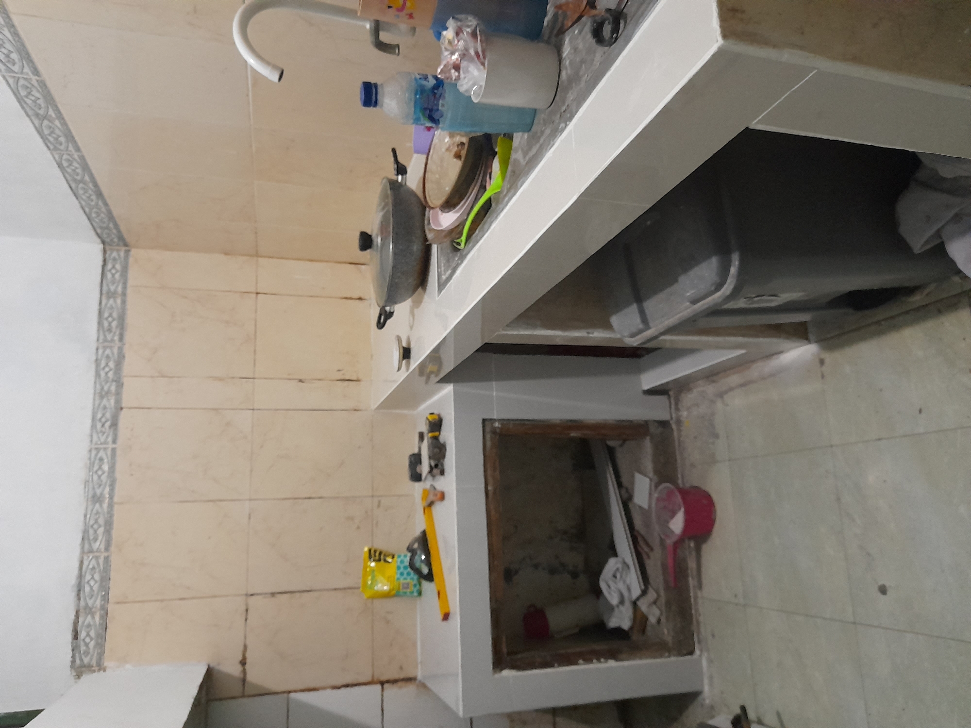 The image depicts a sink and surrounding area in a kitchen or bathroom setting. The sink is cluttered with various items, including a pot with a lid, a water bottle, a container holding wrapped items, dishes, and other cleaning supplies. The faucet is visible above the sink. Beneath the sink is an open cabinet space containing various objects, including what appears to be a red plastic scoop, cloth, and possibly other tools or cleaning supplies. The walls and floor are tiled, with a decorative border along the wall. The overall scene suggests a lived-in space, likely a residential kitchen or bathroom. The geographical location is specified as Gelam, Indonesia.