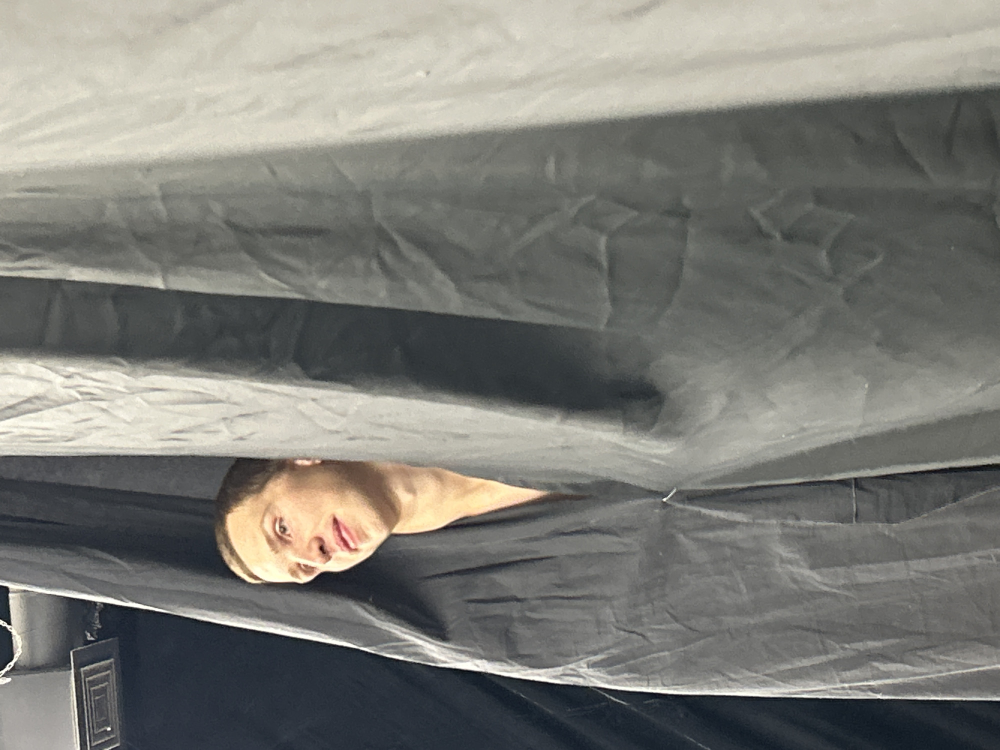 A young adult male with short brown hair and a light complexion is peeking out from behind a dark grey curtain. He is looking to his left with his mouth slightly open. His shoulders and neck are visible, and he appears to be wearing a dark garment that is open at the neck. The background consists of more dark curtains and a hint of what appears to be a light fixture and a vent on the ceiling. The lighting is dim.