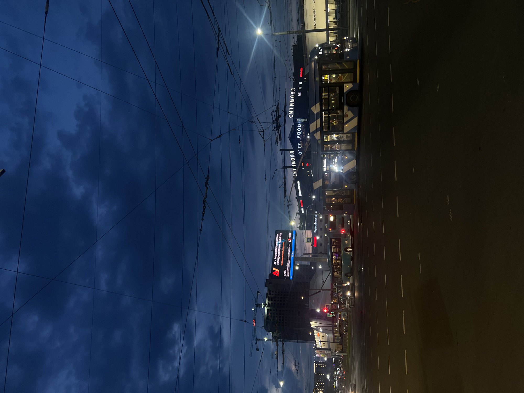 This image depicts an urban street scene at dusk or night, characterized by a dark blue, cloudy sky interwoven with numerous overhead electrical wires. The location is Kolomyazhsky Prospekt 17, Saint Petersburg, Russia, identifiable by the illuminated "СИТИМОЛЛ" (CITIMALL) sign on a prominent commercial building.
In the mid-ground, a multi-lane asphalt road occupies the lower portion of the frame, marked with dashed white lane lines. Traffic is present, with several vehicles visible. A prominent blue and white striped trolleybus, possibly a trolleybus due to the overhead wiring, is positioned in the right-center, facing right. Its front headlights and internal lights are illuminated. A second, lighter-colored trolleybus/bus is visible further back, behind the first one, also illuminated. A white sedan is stopped behind the first bus, with its brake lights illuminated, at a red traffic light on the left side of the road. Other vehicle lights are discernible further down the street.
To the right, a large commercial building features brightly lit signs, including "СИТИМОЛЛ" (CITIMALL), "CITY FOOD", and "СТРОЙМАТЕРИАЛЫ" (BUILDING MATERIALS). On the left side of the street, a multi-story building under construction is visible, topped by two red construction cranes with their booms extended. The building itself shows internal or work lighting. Between the construction site and the commercial building, a large digital billboard displays Russian text: "A7 - РОССИЙСКАЯ СИСТЕМА МЕЖДУНАРОДНЫХ РАСЧЕТОВ" (A7 - RUSSIAN SYSTEM OF INTERNATIONAL SETTLEMENTS) and "A7.RU" with a distinctive red A7 logo. Behind this billboard, additional commercial establishments with illuminated signs, including "Перекресток Супермаркет" (Perekrestok Supermarket), are present. Streetlights throughout the scene are illuminated, casting bright, localized light. No individually discernible people are visible.