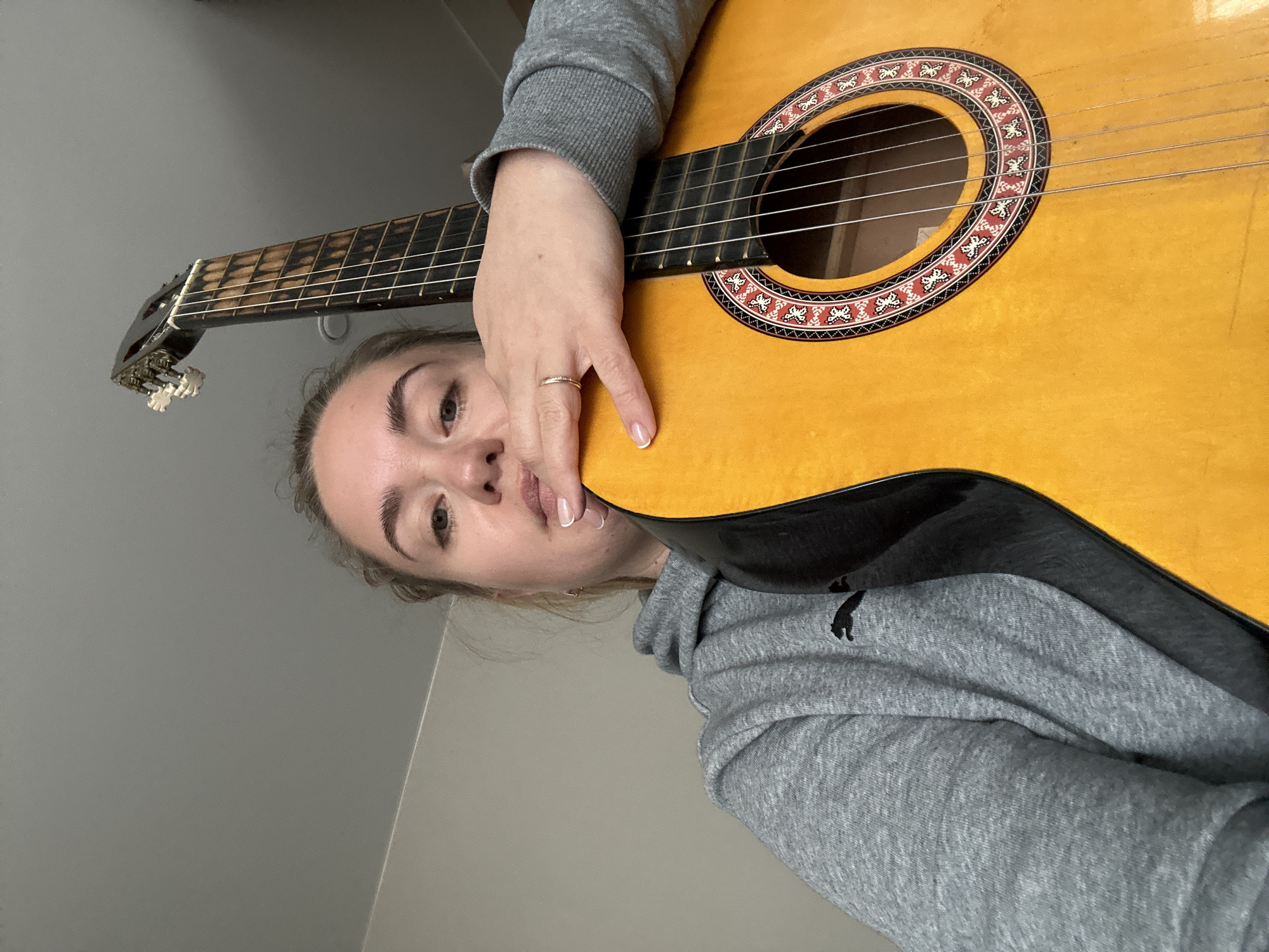 A person is holding a guitar, likely in an indoor setting. The person is wearing a gray hooded sweatshirt. The guitar is a classical guitar with a light-colored top. The location is Уральское, Russia.
