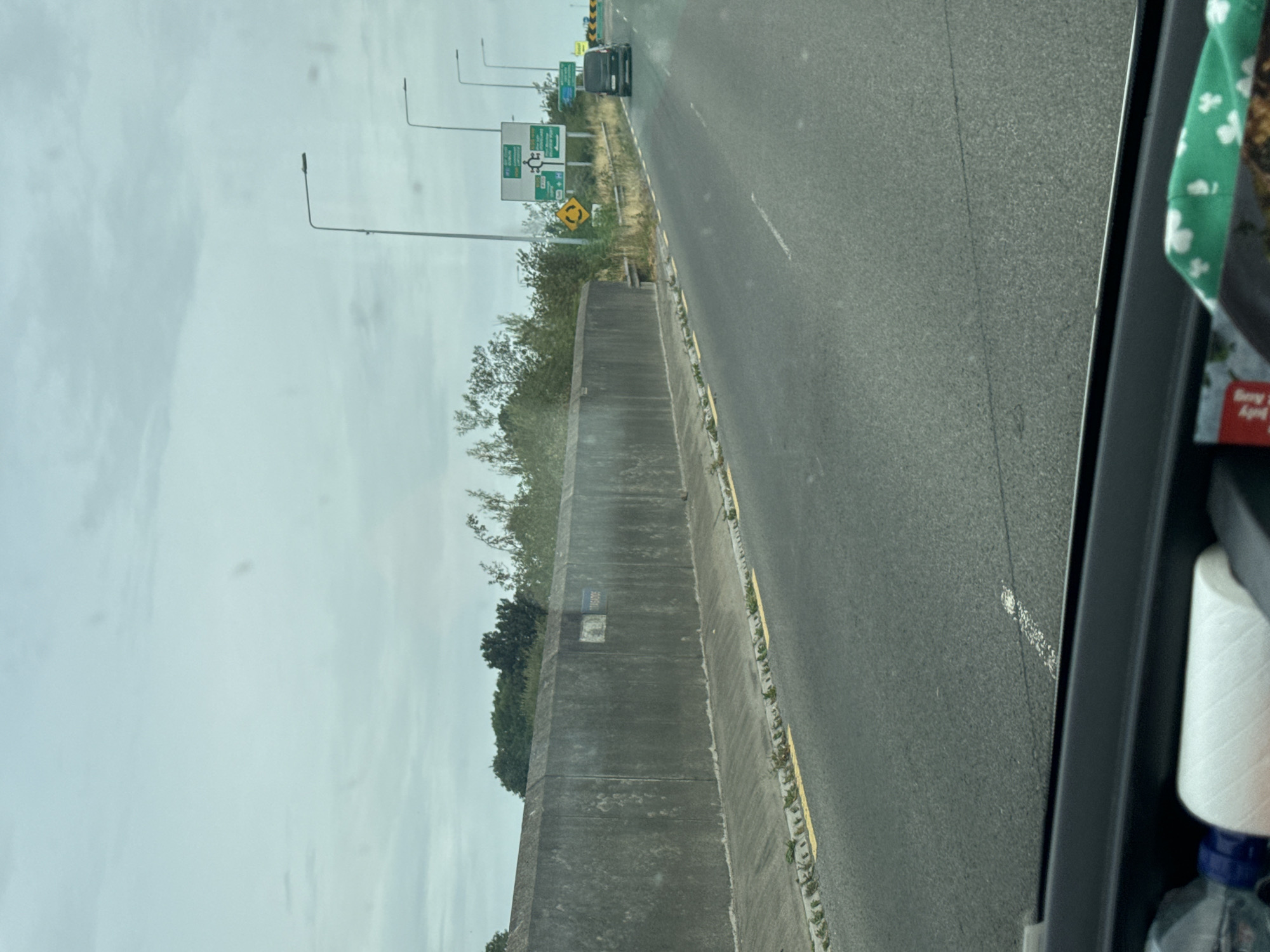The image is a view from a vehicle traveling on a road with a concrete barrier on the left and other vehicles ahead. Overhead, streetlights are visible, and the sky is overcast. On the right side of the road, there are road signs. One sign displays directions to Grannagh, Ireland, with a roundabout symbol and distances to various locations, including the M9 and a hospital. Another sign indicates a roundabout ahead. A dark-colored van is visible in the right lane, and further ahead, there's a yellow safety barrier. The road has white markings and a yellow line along the edge of the barrier. The lower part of the image shows the interior of the vehicle, with a water bottle and a roll of white material in the foreground.