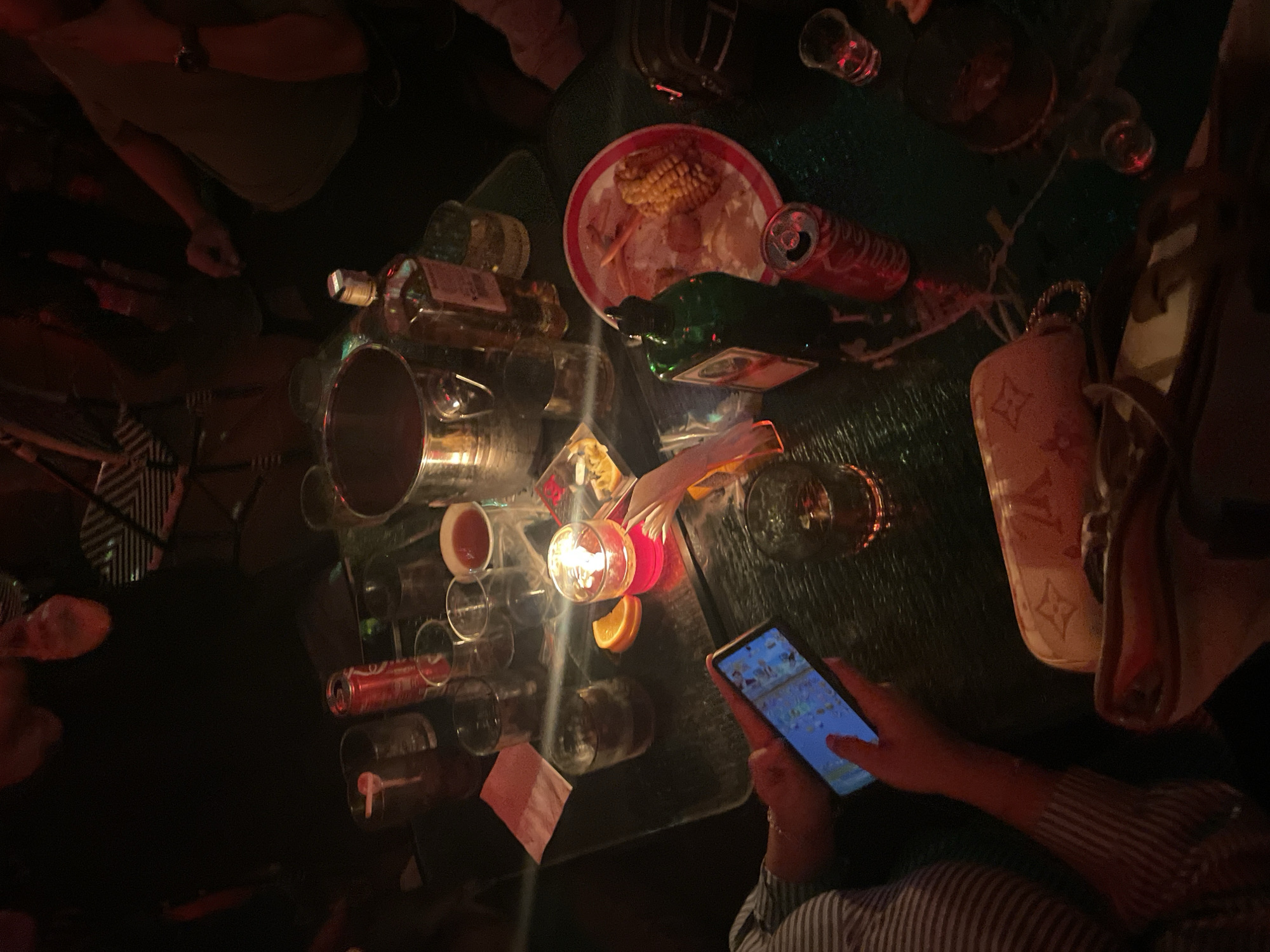 The image captures a dimly lit indoor scene at a venue in Bandung City, Indonesia, likely a bar or lounge, centered around a dark, reflective table. On the table, a metallic ice bucket is positioned alongside an amber-colored liquor bottle and a green bottle resembling Jägermeister. Two red Coca-Cola cans are visible, with one appearing open. Multiple drinking glasses, some containing dark liquid and ice, are dispersed across the table. A red candle in a glass holder illuminates a nearby orange slice and a small dish of yellow snacks. A white plate holds partially consumed food, including a corn on the cob and other remnants. Pink napkins and several small shot glasses are also present. In the left foreground, an individual wearing a striped shirt holds a smartphone with both hands, the screen emitting a blue light. Another person is partially visible in the upper right, wearing a light-colored shirt and a watch on their left wrist. The background is indistinct, showing silhouettes of people and objects, consistent with a low-light social setting.