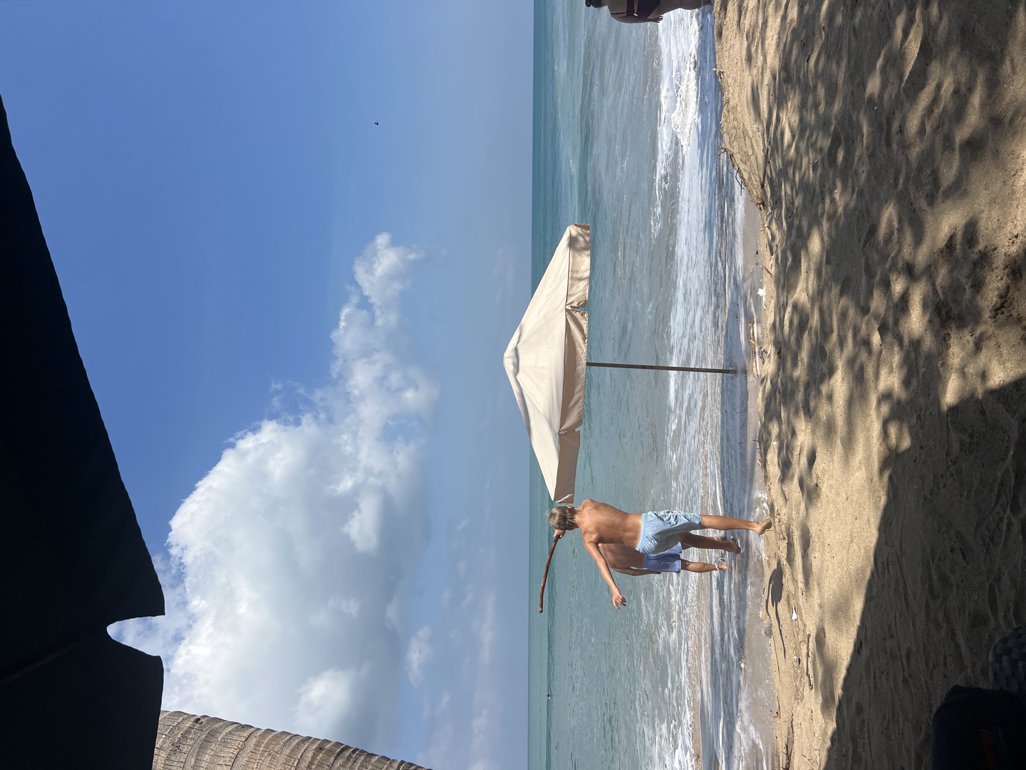 The image depicts a beach scene in Kuta, Indonesia, on a sunny day with a clear blue sky and scattered clouds. In the foreground, two young men are standing in the shallow water at the edge of the beach, facing the ocean. One of the men, closer to the camera and in focus, is wearing light blue swim shorts and is holding a long, thin stick, appearing to be mid-action, possibly about to throw it or pose with it. The other man, slightly further back and less in focus, is wearing darker blue swim shorts.
A large, light-colored beach umbrella is positioned on the sand behind the men, its pole firmly planted in the ground. The ocean extends to the horizon, with gentle waves breaking on the shore. In the distance, on the water, there are small figures that appear to be surfers or swimmers. To the right of the frame, a person, partially visible from the back and wearing a dark-colored bikini, is standing in the water, also facing the ocean.
The beach itself is sandy, with some dry leaves and small pieces of debris scattered on the surface. Shadows from palm trees or other foliage are cast across the sand in the foreground, indicating that the photo was taken from beneath a shaded area. A portion of a palm tree trunk is visible on the far left of the image. The overall impression is one of a relaxed, tropical beach environment.