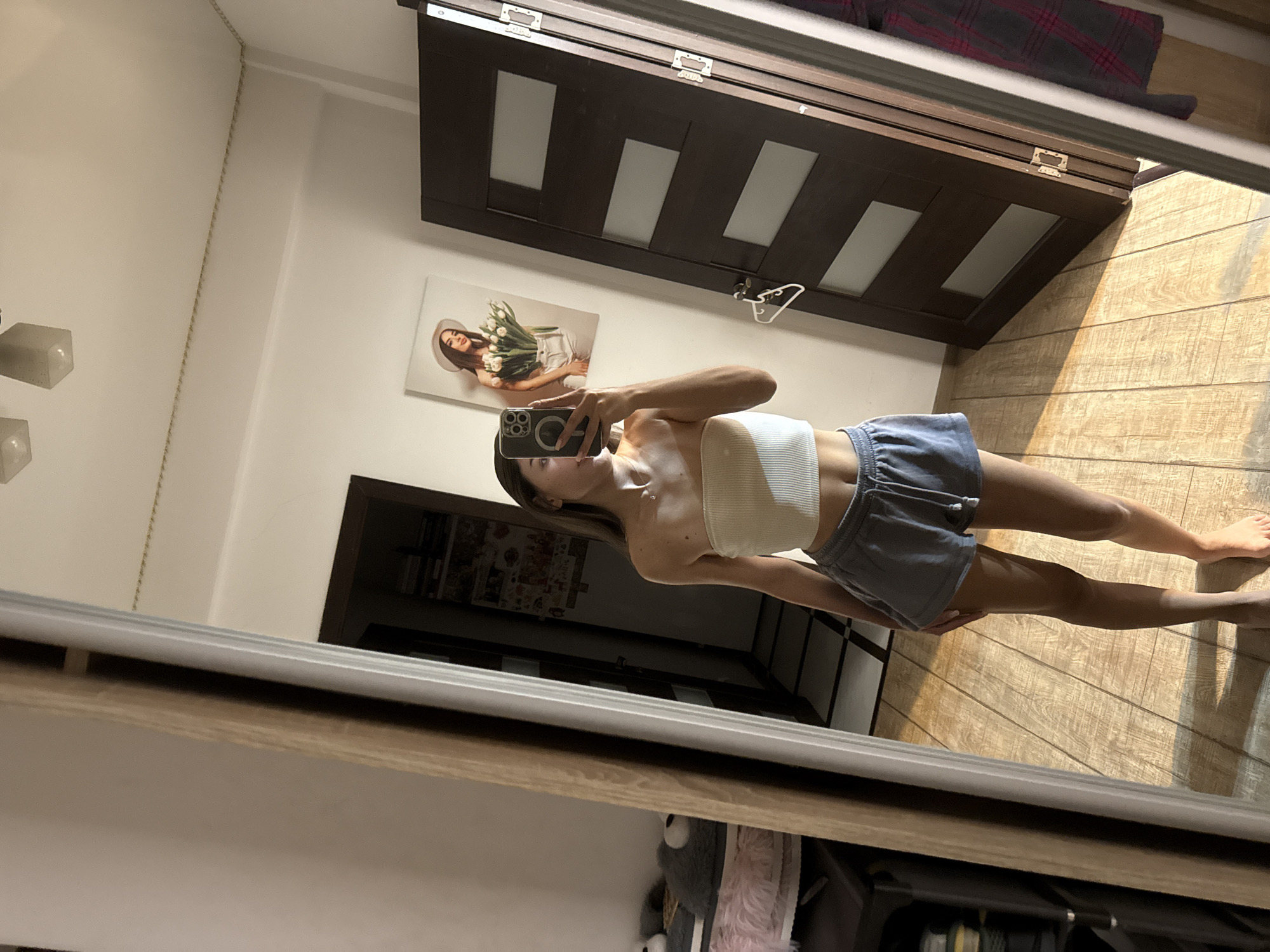 The image is a full-length mirror selfie taken indoors, likely in a bedroom. The subject, a young woman, stands facing the mirror, holding a smartphone to take the picture. She is wearing a light-colored strapless crop top and grey athletic shorts. Her hair is long and brown, and she has minimal jewelry on.
The environment appears to be a room with wooden floors. To the left of the subject, a large wardrobe or closet is partially visible, with some shelves and items on them. A plush grey fuzzy object, possibly a chair cushion or a stuffed animal, is also visible. To the right of the subject, a dark wooden door with glass panels is open. On the wall behind the subject, a framed picture of a woman holding a bouquet of tulips is hanging. Above the picture, two modern ceiling lights are visible. The overall lighting suggests it might be daytime, with sunlight casting shadows on the floor. The scene does not provide any specific indicators to determine its location within the city of Chișinău, Moldova.
