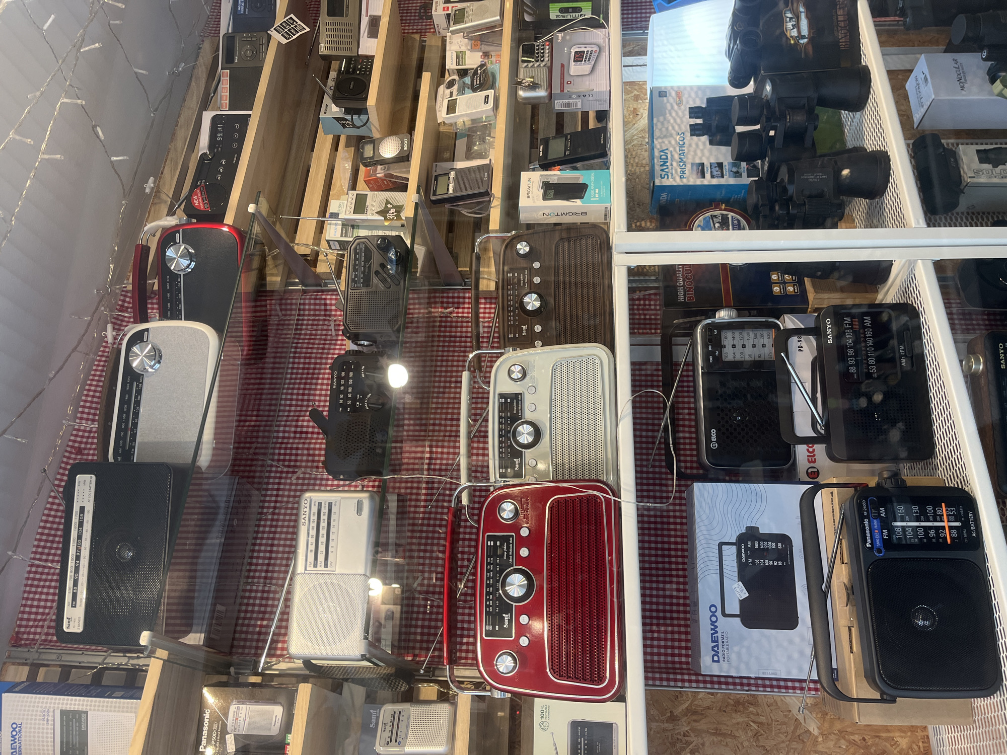 The image shows a shop window display in A Coruña, Spain. The main focus is on a collection of radios, both vintage and modern, arranged on shelves. Some radios are in boxes, displaying brand names like Daewoo, Panasonic, Sanyo, Elco and Sami. Also, there are the name Brightgon and Muse on them. Many of the radios are red, white, black or grey, with both analog and digital controls. Some of the radios have extended antennas. Boxes containing binoculars and monoculars are also visible in the upper right section of the display. There are no people visible in the frame. The radios are arranged against a backdrop of red and white checkered material. Strings of lights are placed behind the material.