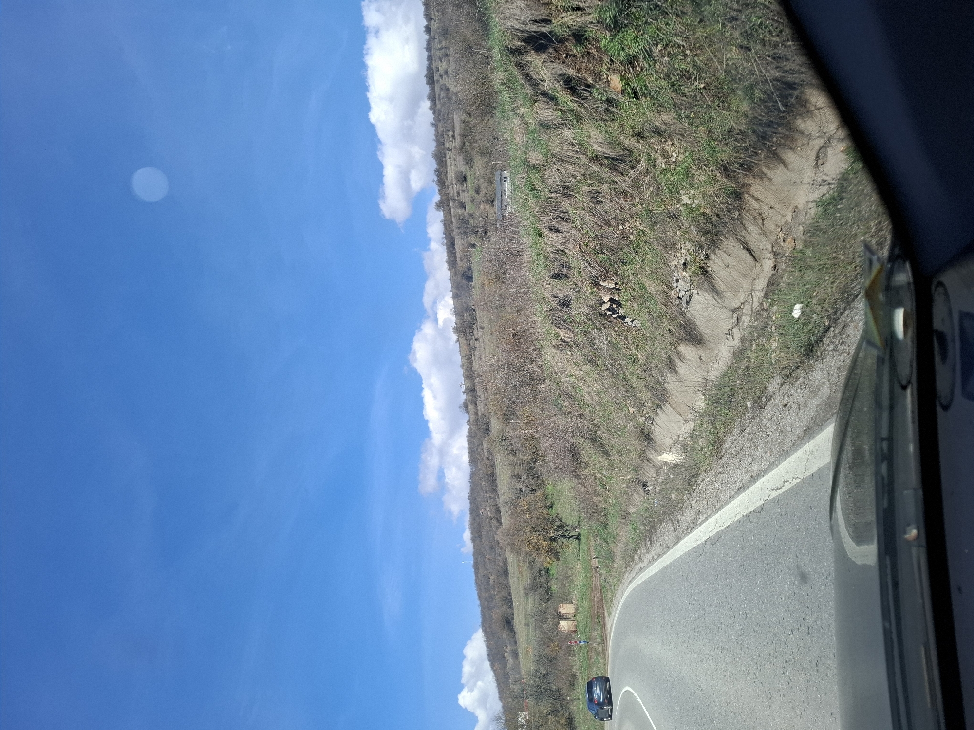 The image shows a view from inside a vehicle on a road in Iskrets, Bulgaria. The road curves to the right, marked by a white line on the shoulder. A dark-colored car is visible on the road ahead. The landscape to the right of the road is characterized by dry vegetation and a slight slope. In the distance, there's a hill partially covered with trees and shrubs. The sky is blue with some white clouds. A bright spot, possibly glare from the sun, is visible in the upper left corner of the image.