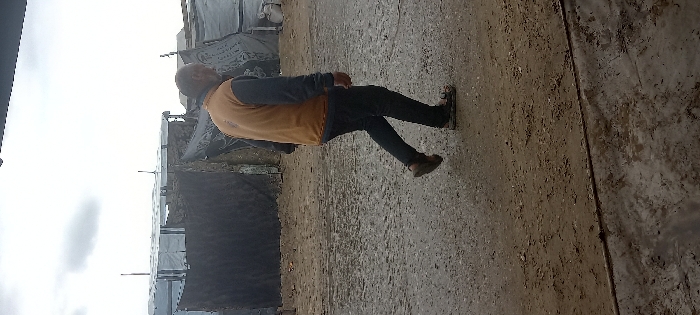 The image depicts an outdoor scene in Khan Yunis, Palestinian Territory. An adult male, appearing middle-aged, is shown walking from right to left across a mixed ground surface. He wears a mustard yellow sleeveless vest over a dark long-sleeved shirt, dark trousers, and sandals; his right foot is elevated in mid-stride. The ground beneath him consists of damp, light brown packed earth on the left, transitioning to a darker, wet grey concrete or paved area on the right, with a clear dividing line between the two surfaces. In the background, several improvised structures are visible, composed of various materials including dark and light-colored tarpaulins draped over frames, with slender support poles extending upwards. The sky overhead is uniformly overcast and light grey. No other individuals or immediate interactions are observable.
