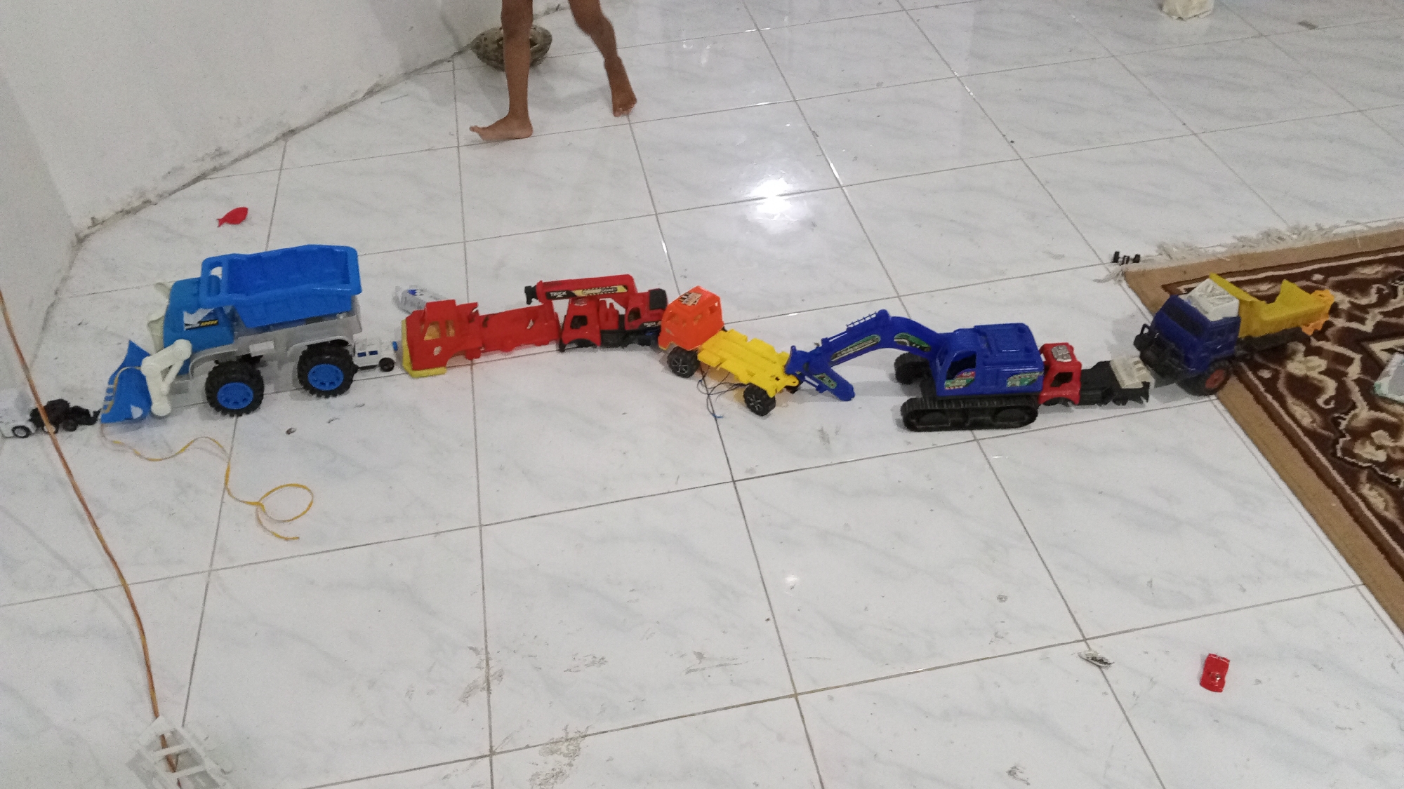 In Lhokseumawe, Indonesia, a child's legs are visible at the top of the frame, suggesting a child is playing. They are on a tiled floor with a white and gray checkered pattern. A collection of colorful toy vehicles is arranged in a line, appearing to be connected or engaged in some form of play.
The toys include:
* A blue and gray front-loader truck on the left, with a yellow string attached to it, leading out of frame.
* Several red toy trucks, some with "TRUCK" printed on them.
* An orange truck with a yellow trailer.
* A blue excavator with black tracks and a yellow bucket.
* A blue truck with a yellow trailer on the far right, partially on a patterned rug.
There are also a few smaller scattered red and dark-colored toy pieces on the floor. The lighting suggests an indoor environment, possibly during the day. The overall scene depicts a child's playtime activity with toys.