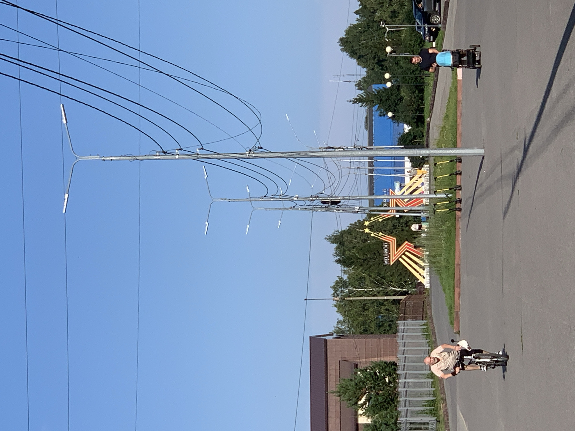 The image shows a man riding a bicycle down a paved road in Prokopyevsk, Russia. In the background, there are several streetlights, a building, trees, and a monument. To the right of the monument, another man is pushing a stroller. The sky is clear and blue. The monument appears to be a star with the word "ПОБЕДЫ" written on it, which translates to "Victory".