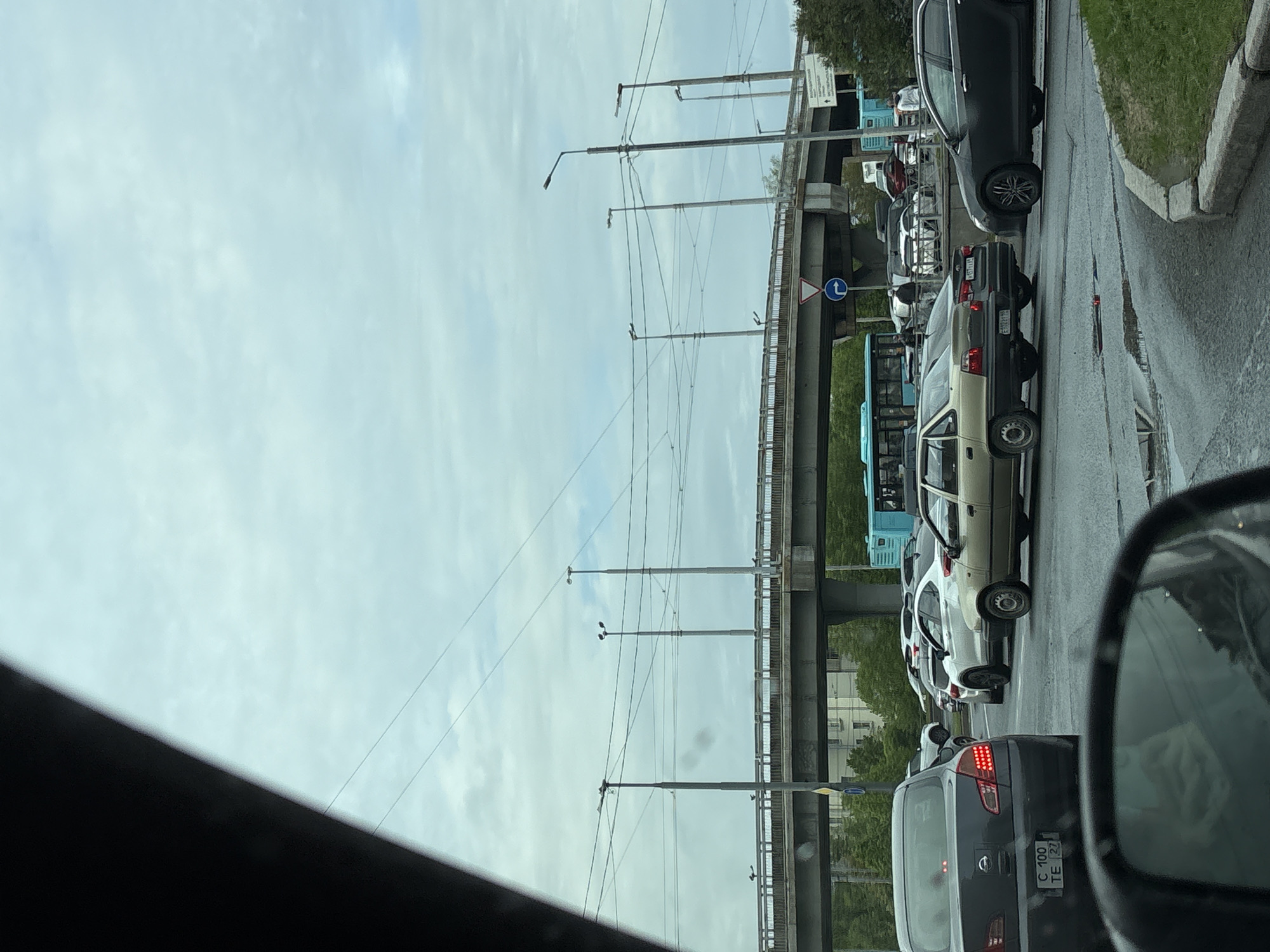 The image shows a traffic jam on a city street in Saint Petersburg, Russia, with cars lined up bumper-to-bumper. An elevated roadway or bridge is visible overhead. The weather appears overcast, and there are puddles on the road, indicating recent rain. Streetlights and power lines run along the road. A blue and white traffic sign is present. The license plate of the car closest to the camera includes the Cyrillic letter "C" and the numbers "100" and "27." The scene is observed from inside another vehicle, as evidenced by the partially visible rearview mirror and the car's interior.