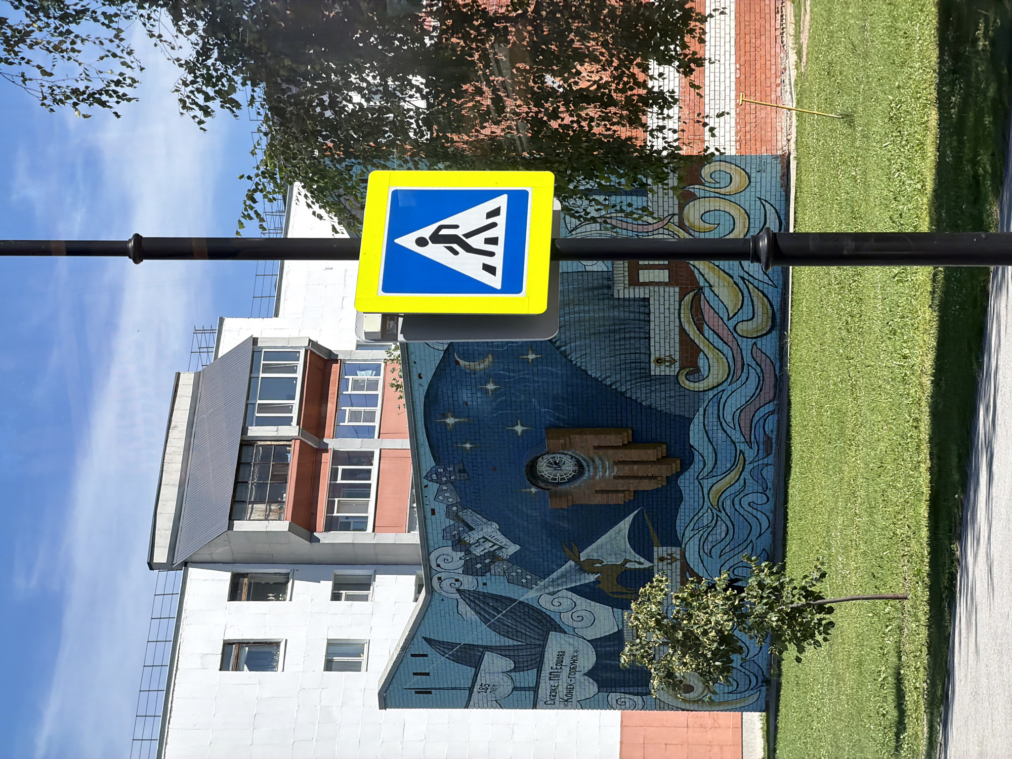 The image depicts an outdoor scene with a pedestrian crossing sign in the foreground, mounted on a dark pole. The sign is square with a yellow border and a blue interior, featuring a white silhouette of a person walking. Behind the sign, a large, colorful mosaic mural covers the side of a building. The mural is abstract with swirling blue and yellow patterns, and also includes depictions of ships, stars, and possibly a clock face.
To the left of the mural, part of a multi-story building is visible, with a facade of white and reddish-brown sections, and numerous windows. To the right of the mural, a neatly manicured green lawn stretches out, with a paved pathway running alongside it. A small, leafy tree grows in front of the mural. In the upper portion of the image, dappled sunlight filters through the leaves of a large tree, casting shadows. The sky is blue with some scattered clouds.
The activity taking place is not explicitly shown, but the presence of the pedestrian crossing sign suggests a public area where people might be walking or crossing a street. The bright sunlight and blue sky indicate it is daytime, likely a clear and pleasant day.
There is text visible on the mural, in the lower right quadrant. It appears to be in Russian. Part of it reads "Сказке П. П. Ершова «Конек-Горбунок»", which translates to "Fairytale by P. P. Ershov 'The Little Humpbacked Horse'". Another smaller inscription says "185 лет" meaning "185 years". The overall mood is bright and slightly whimsical due to the mural.