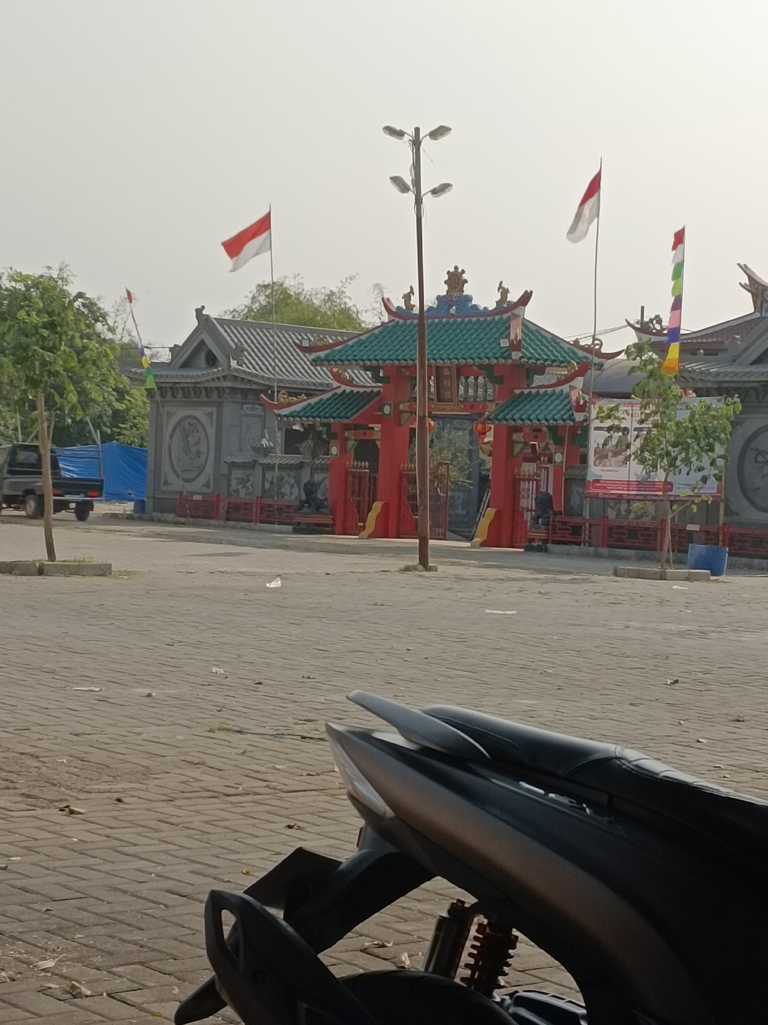 This image shows a street scene with a prominent red and green pagoda-style structure in the background, likely a temple or cultural site. Two Indonesian flags are visible, one red and white and another with multiple colors, suggesting a national celebration or event. In the foreground, the rear section of a black motorcycle is visible. The ground is paved with brick. A black pickup truck is parked on the left side of the image, and a blue tarp is draped over something behind it. The sky is overcast. The presence of the Indonesian flags and the architectural style of the building suggest the location is Karawang, Indonesia, possibly a public space or cultural landmark.