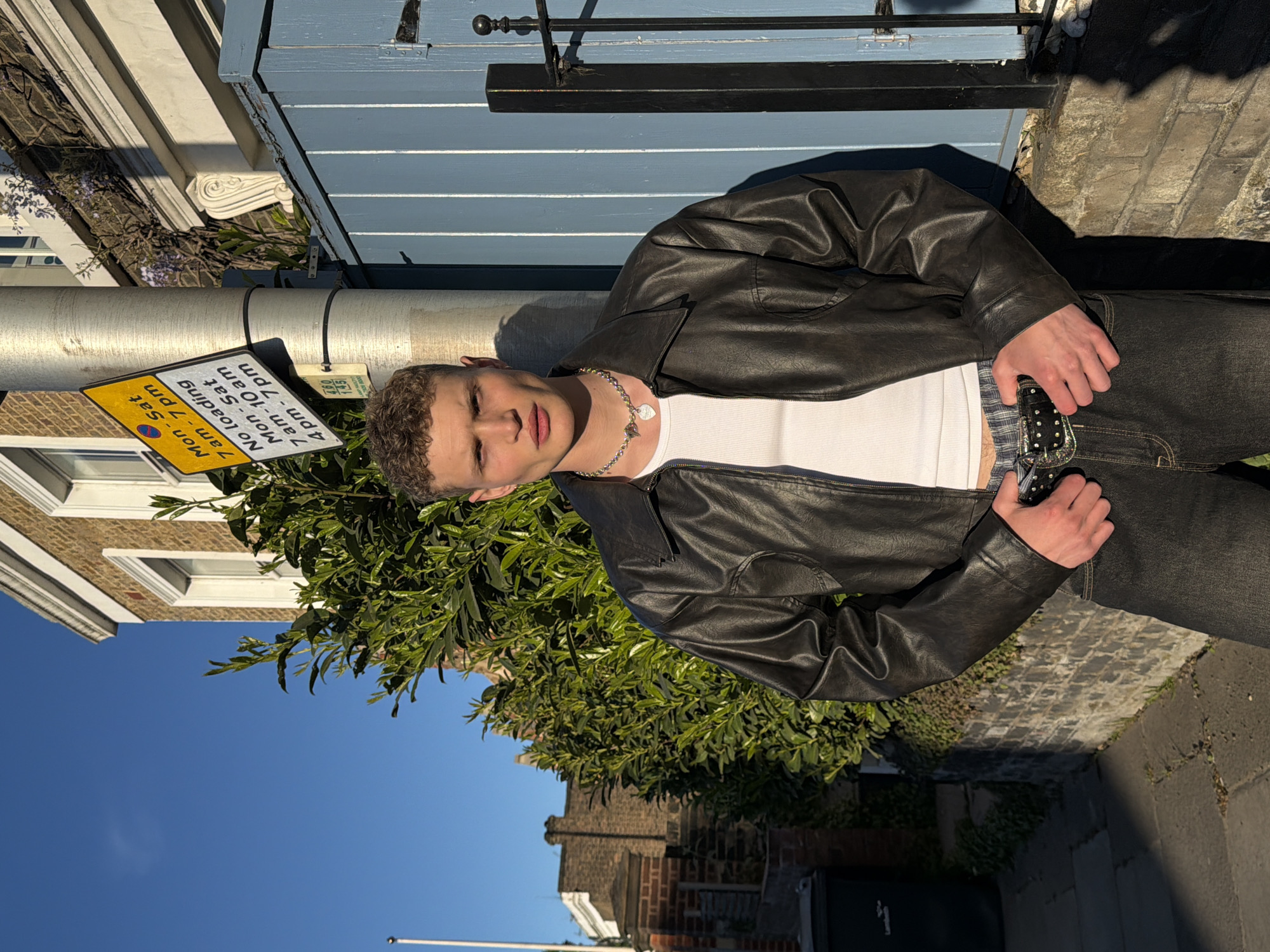 A young man poses outdoors in London, United Kingdom, wearing a black leather jacket, a white ribbed tank top, and dark jeans with a decorative black belt. He stands close to a building with a light blue door or shutters and ornate white details above. A street sign attached to a silver pole shows "No loading" restrictions, with specific times listed for Monday to Saturday. Green foliage partially obscures the view behind the man. Buildings and sky are visible in the background. The image is taken at an angle, rotated for a different perspective. The surface on which the man stands appears to be paved.