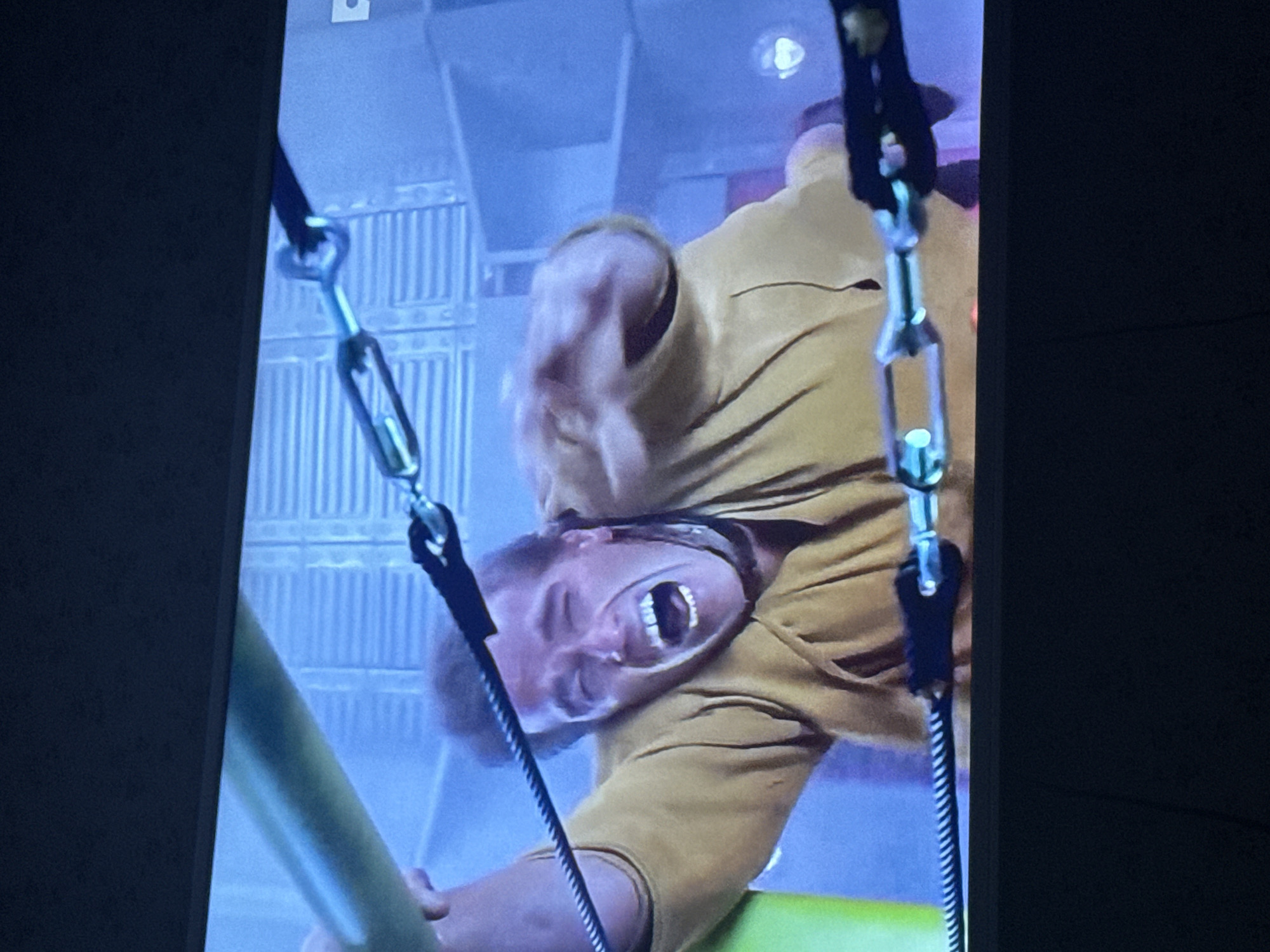 A man wearing a yellow shirt is suspended upside down by cables and metal turnbuckles. He is grimacing, and his mouth is open. A green cylindrical object is near his left arm. The background features a metal grate wall. The image appears to be a still from a video or movie displayed on a screen. No evidence indicates a location in Mytishchi, Russia.