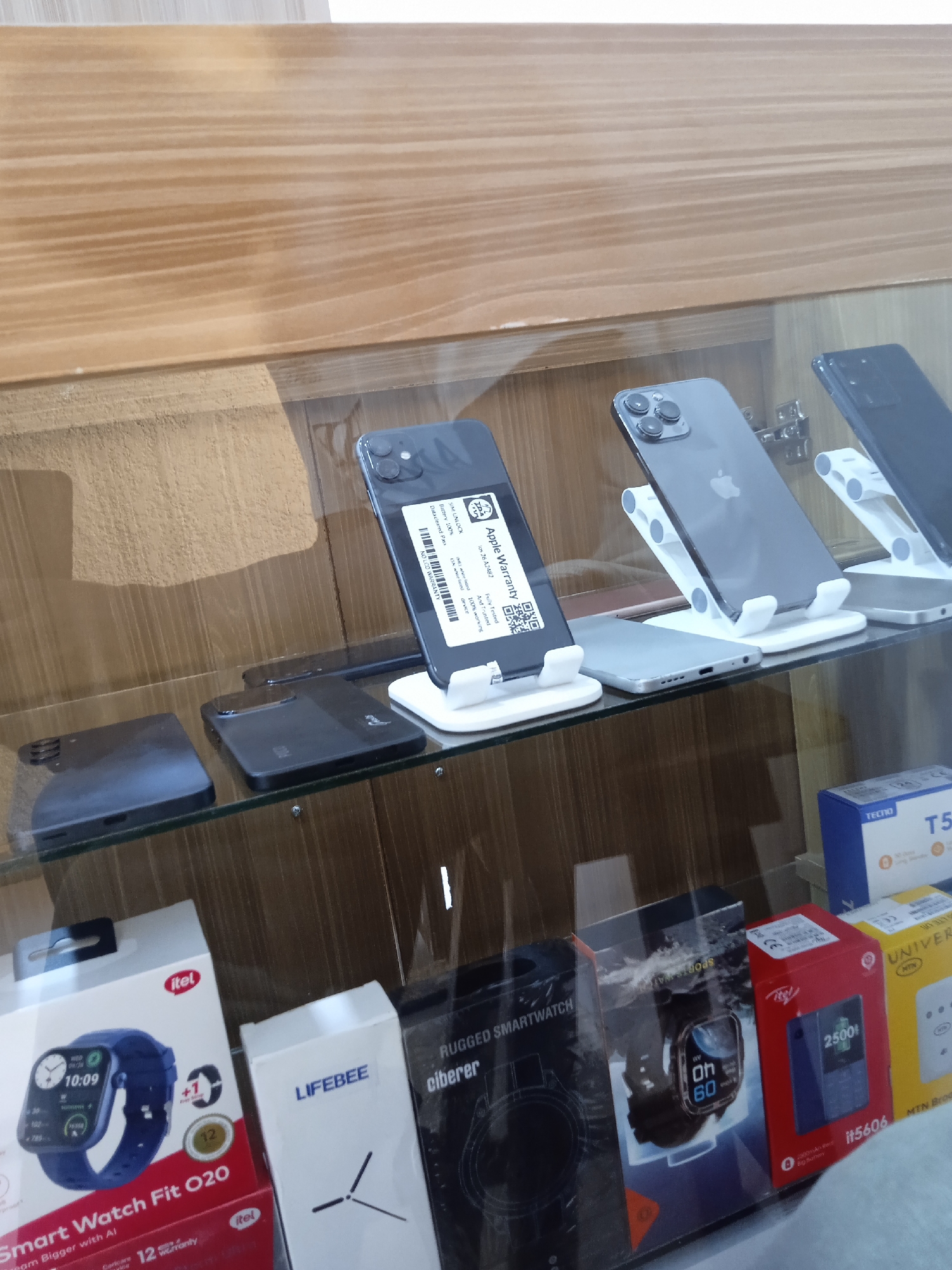 The image shows a display case in a shop, likely located in Jos, Nigeria, based on the brands and products present. The display features mobile phones and smartwatches, arranged on two shelves within the glass-fronted case.
On the top shelf, there are several smartphones, some of which are displayed on small white stands. These include what appear to be Apple iPhones and other models. One of the phones has a label indicating "Apple Warranty", mentioning "Datacleared Pass," "Battery 100%," and "SIM UNLOCK."
The lower shelf contains the retail packaging for various smartwatches and mobile phones. Brands like "itel" and "TECNO" are visible, common in the Nigerian market. There are also boxes for "LIFEBEE" and "ciberer" rugged smartwatches.
The environment appears to be a retail setting, with a clean and organized display. The lighting is somewhat diffused, casting reflections on the glass case. The backdrop is a plain wooden surface.
