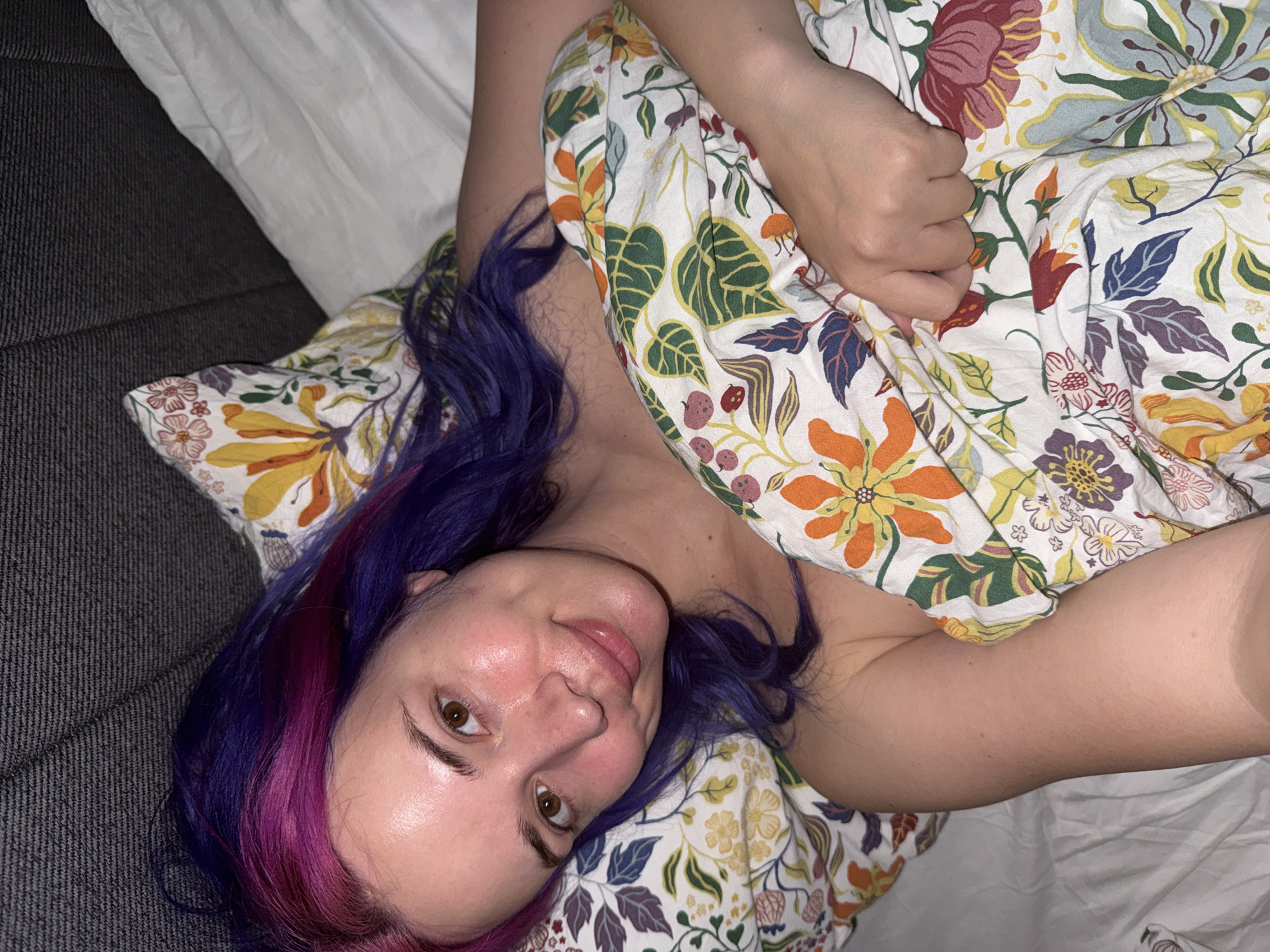 An overhead indoor shot captures a woman lying in a relaxed position in the City of Zagreb, Croatia. She looks directly at the camera with a gentle, natural expression and a subtle smile. Her long hair, strikingly dyed in shades of purple and pink, spreads around her head. Her face shows a clear complexion with light brown eyes.
The woman is partially covered by a vibrant floral blanket, which features a cheerful pattern of various flowers and leaves in red, orange, yellow, green, and blue on a white background. One of her bare arms is visible, resting on the blanket.
The background consists of a white sheet or pillow near her head, and to her left, a dark grey, textured fabric surface, possibly a couch or bed frame. The indoor lighting appears soft, suggesting an evening or nighttime setting. The scene conveys a sense of comfort and informality. No visible text is present in the image.