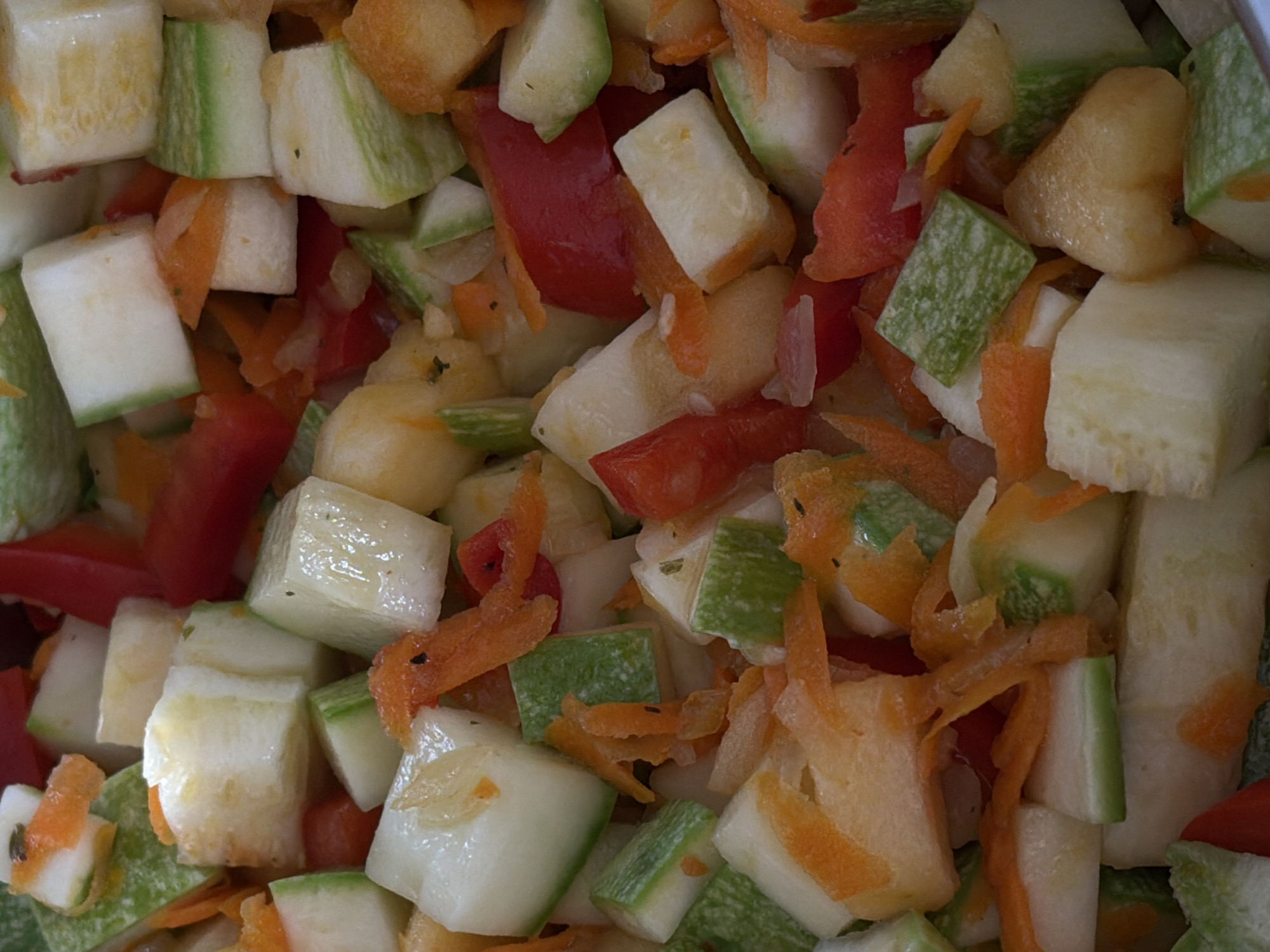 The image is a close-up, overhead shot of a mixture of chopped vegetables, primarily zucchini and carrots, with some red bell pepper pieces. The vegetables are cut into roughly uniform, bite-sized cubes and strips, appearing to be raw or lightly blanched. Some pieces show the distinct green skin of zucchini, while others are the pale flesh. The carrots are shredded or julienned and are bright orange. Red bell pepper pieces are scattered throughout, providing pops of color. The overall impression is of ingredients prepared for a dish, possibly a stir-fry, salad, or soup. There is no indication of any specific location within Khmelnytskyi, Ukraine, nor are there any people, actions, or interactions depicted. The scene is a still life of raw food preparation.