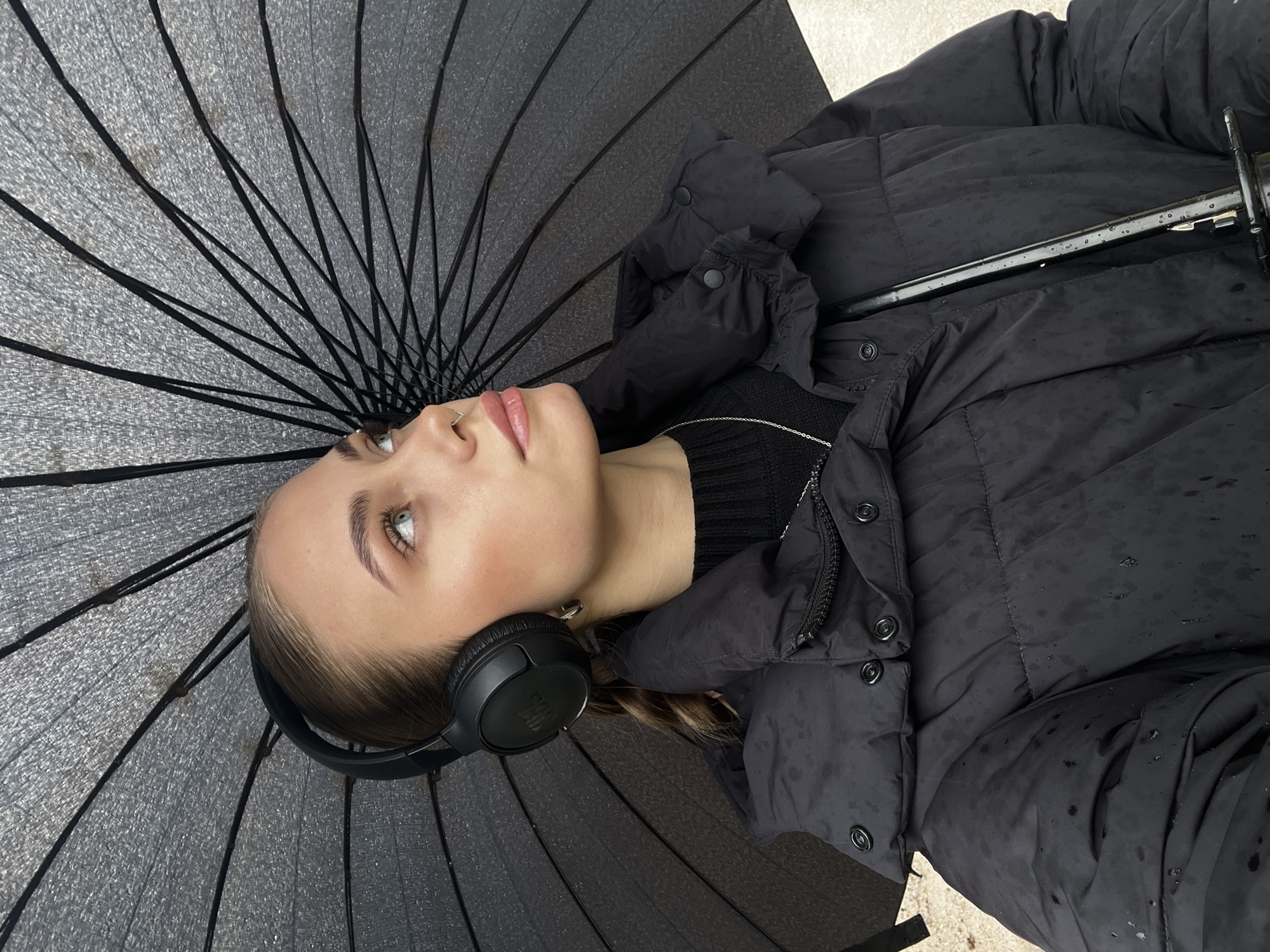 A young woman is depicted lying outdoors, looking upwards, with a large black umbrella partially open above her. She is wearing a black puffy jacket over a black ribbed sweater, and black over-ear headphones with "JBL" visible on the left earcup. A silver chain necklace and a small nose ring adorn her. Water droplets are scattered across her jacket, indicating recent or ongoing rain. Her expression is calm and contemplative as she gazes upwards. The underside of the umbrella, with its dark ribs and textured dark grey fabric, fills a significant portion of the frame, providing shade. The background beyond the umbrella is lightly visible in the top right and bottom left, suggesting an outdoor, possibly paved area. The image was captured in Фоки, Russia, during daytime.
