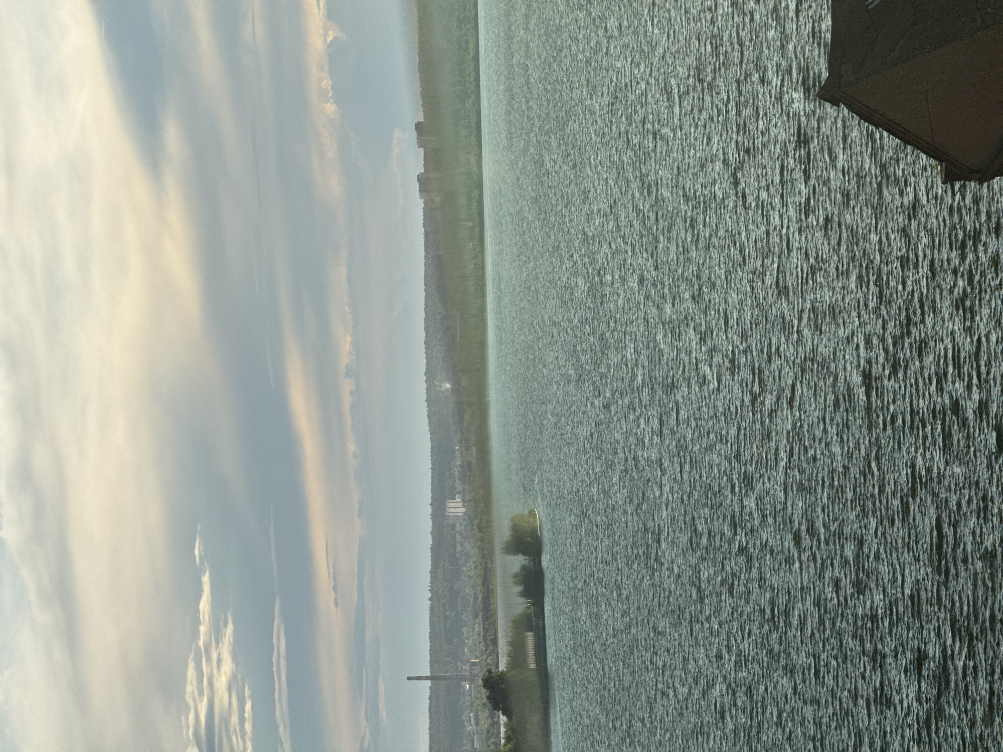 The image depicts a body of water, likely the Izhevsk Pond, with a view towards the city of Izhevsk, Russia. The water surface is textured with numerous small waves. The opposite shore features a line of trees and buildings, including a tall chimney stack. The sky is partly cloudy, with some areas of lighter, yellowish illumination. A dark, angled object is visible in the upper right corner, possibly a portion of a building or structure. The photograph is taken from an elevated position.