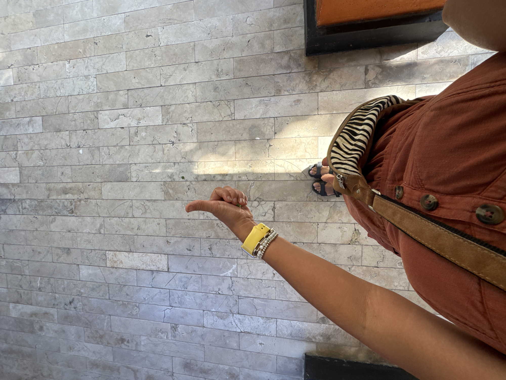 The image shows a first-person perspective of a person standing on a tiled floor, possibly in Kuta Selatan, Indonesia. The person is wearing an orange jumpsuit, black sandals, and has a zebra-print bag with a brown strap slung over their shoulder. They are giving a thumbs-up with their left hand, which is adorned with several bracelets, including a bright yellow one. Sunlight casts shadows on the tiled floor, which is made of rectangular, light-colored stones. To the right, a dark structure with an orange accent is visible.