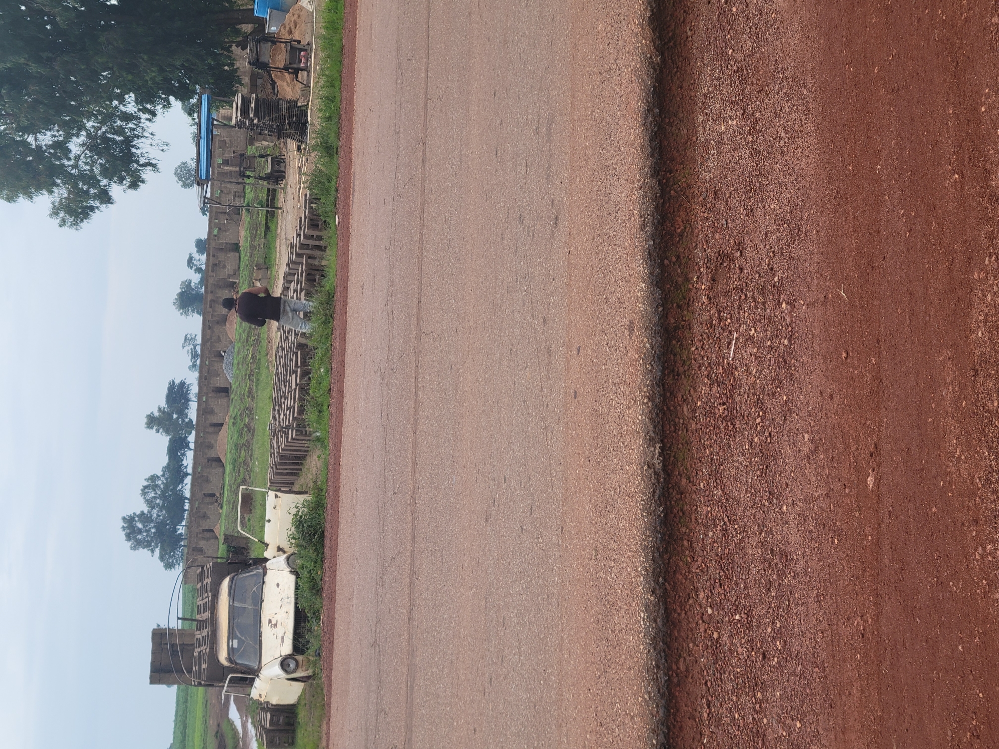The image shows an outdoor scene with an abandoned car, piles of bricks, and unfinished buildings in the background. A man is standing near some bricks, looking away from the camera. The ground in the foreground is a mix of asphalt and dirt. The overall impression is one of a construction site or a rural area where building materials are being manufactured or stored. The sky is overcast.