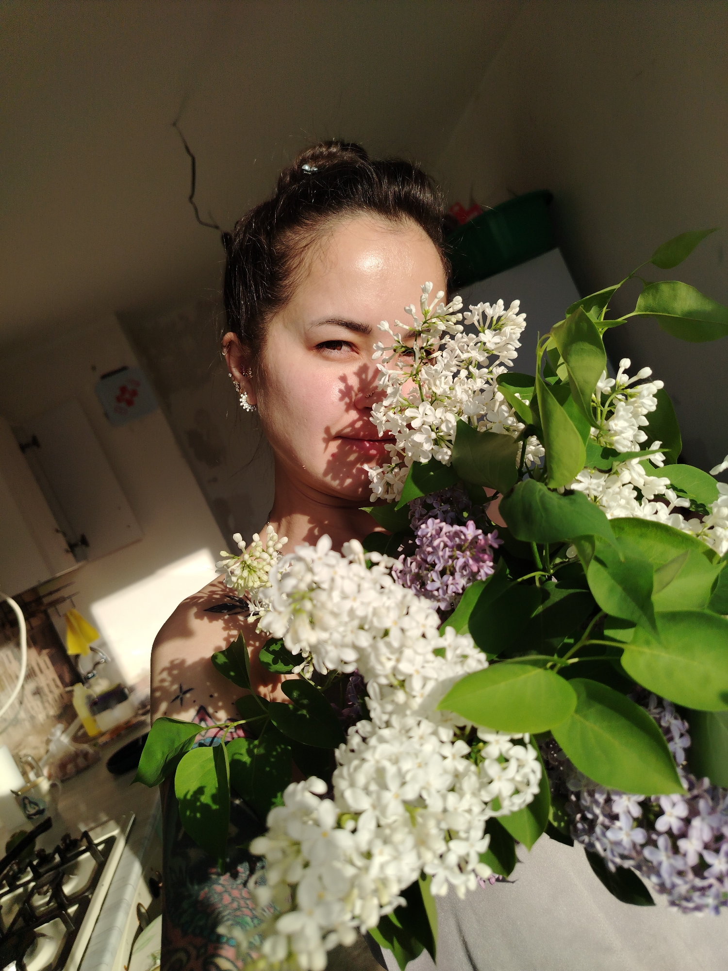 A woman is holding a bouquet of lilac flowers, partially obscuring her face. She is in a room with white walls and cabinets. A stove is visible in the foreground on the left. A crack is visible in the ceiling. The photograph was taken indoors in the city of Noviy, Russia.