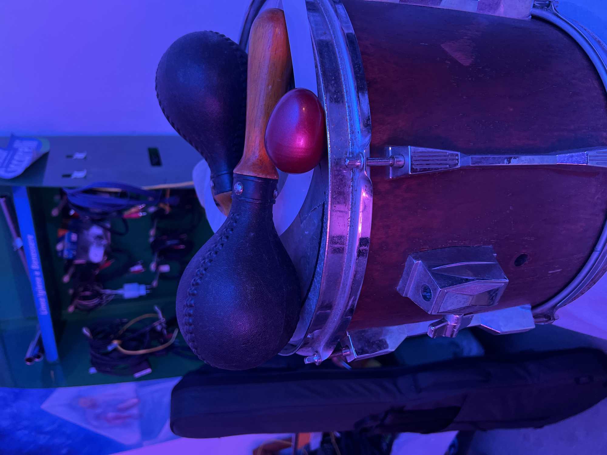 This image, captured in Santeramo in Colle, Italy, presents an indoor scene under pronounced artificial blue and purple lighting. The foreground is dominated by a dark reddish-brown tom drum, featuring worn chrome hardware including lugs and a rim. Resting on the drum's head are three percussion instruments: two large maracas with dark, textured (likely leather) heads and wooden handles, one positioned slightly behind the other; and a smaller, glossy, reddish-purple egg-shaped shaker placed prominently in front of the maracas.
To the left of the drum, a dark, vertical object consistent with a soft guitar case is partially visible. Further left and higher, a light blue wall section features a framed image or poster depicting a figure with reddish tones against a lighter background.
The background centralizes on a dark green wall or panel. A blue horizontal shelf or sign is mounted on this panel, displaying two silver, cylindrical objects. Below this shelf, a cluster of tangled cables in black, yellow, white, and red are hanging. Above the blue shelf, partially visible white text spells "Streelli" or "Streeli". Directly below the blue shelf, white Italian text on the green panel reads "Linea Igiene e Benessere" (Hygiene and Wellness Line). To the right of the green panel, a plain wall exhibits a consistent light purple-blue color. The entire environment is saturated with blue and purple light, casting vibrant reflections on metallic surfaces. No individuals are present in the scene.