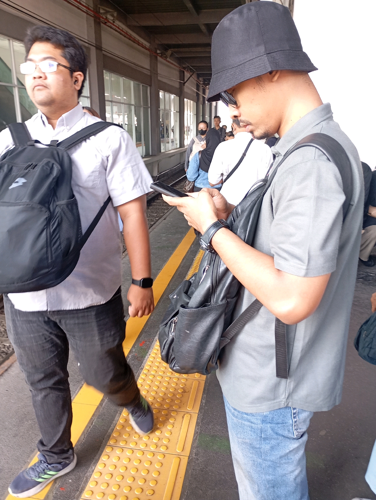 The image shows two men on a train platform in West Jakarta, Indonesia. The man on the left is wearing a light-colored collared shirt, dark pants, glasses, and a black backpack. He is looking to his left and appears to be walking. The man on the right is wearing a grey polo shirt, blue jeans, sunglasses, and a black bucket hat. He is holding a smartphone in his hands and looking down at it. He is also wearing a black backpack. The platform has yellow tactile paving and a yellow line marking the edge of the platform. In the background, other people can be seen waiting for a train, and there is a covered walkway structure overhead.