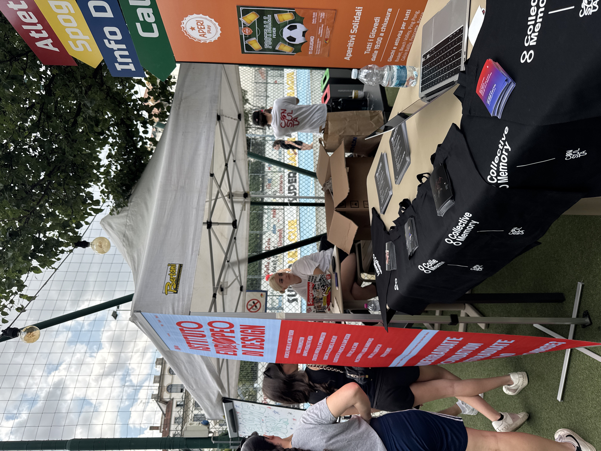 The image captures an outdoor event set on artificial green turf in Milan, Italy. A prominent white canopy tent, bearing a "Bertoni" logo on its canopy, occupies the mid-ground.
Under this tent, to the left, a red vertical banner advertises the "ISTITUTO EUROPEO DI DESIGN," detailing various programs such as INTERIOR & PRODUCT & MOBILITY DESIGN, FASHION, GRAPHIC & INTERACTIVE MEDIA, and others. To the right of the banner, a woman with blonde hair, wearing a white t-shirt with "Supreme" visible, is seated at a desk, looking to her right. Behind her, another individual in a white t-shirt with "CON SUL LA" on the back faces away from the camera, standing near several open cardboard boxes.
In the foreground, two women are partially visible on the left. One wears a grey t-shirt and dark blue shorts, while the other wears a black top and black shorts. Both appear to be observing the booth area.
To the far right, a tall orange vertical sign features an "APERI Play" logo and advertises "FOOTBALL FEVER" on "11 GIUGNO" (June 11) from 19:00. Below this, "Aperitivi Solidali" (Solidarity Aperitifs) are promoted for "Tutti i Giovedì dalle 19:00 a chiusura" (Every Thursday from 7 PM until closing), listing activities such as Basket, Beach Volley, Ping Pong, Calcio Balilla, Freccette, and Bocce. Above the orange sign, multi-colored directional arrows indicate "Atleta" (red), "Spog" (yellow, likely Spogliatoi for changing rooms), "Info D" (blue), and "Cal" (green, likely Calcio for football).
On the tables in the foreground and under the tent, black tablecloths are draped, bearing a white "Collective Memory" logo and accompanying text. Displayed on these tables are multiple black tote bags and small brochures with red and blue designs, also featuring the "Collective Memory" branding. A silver laptop is open on one table, next to which rests a bottle of Acqua Dolomia mineral water.
Overhead, string lights with visible bulbs are suspended. The background features a green mesh fence, a light blue surface (possibly a sports court), and distant buildings showcasing traditional architectural styles under a partly cloudy sky.