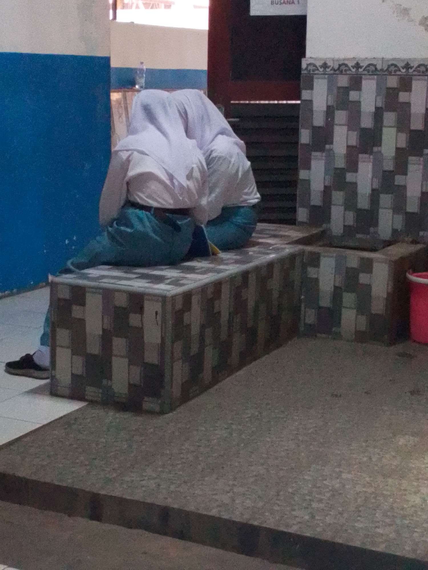 Two individuals, appearing to be students wearing white hijabs and light blue uniforms, are seated on a tiled bench. One is hunched over with their head down, possibly sleeping or resting. The other is leaning against them. The bench is situated in an indoor environment with tiled walls and a blue wall to the left. A red bucket is visible to the right of the bench. The scene suggests a school or public building setting in Probolinggo, Indonesia, possibly a waiting area or corridor.