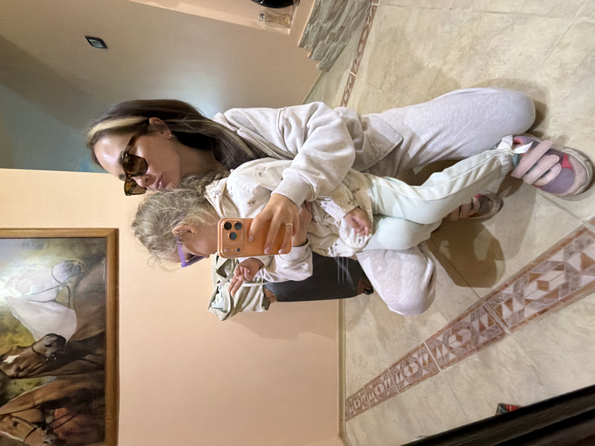A person wearing sunglasses, a light-colored sweatshirt, and sweatpants is sitting on a tile floor holding a young child. The child is wearing light-colored pants and a patterned shirt. The person is holding a cell phone up, possibly taking a picture or recording a video. There is a framed horse painting on the wall to the left. The location is Kamyshin, Russia.