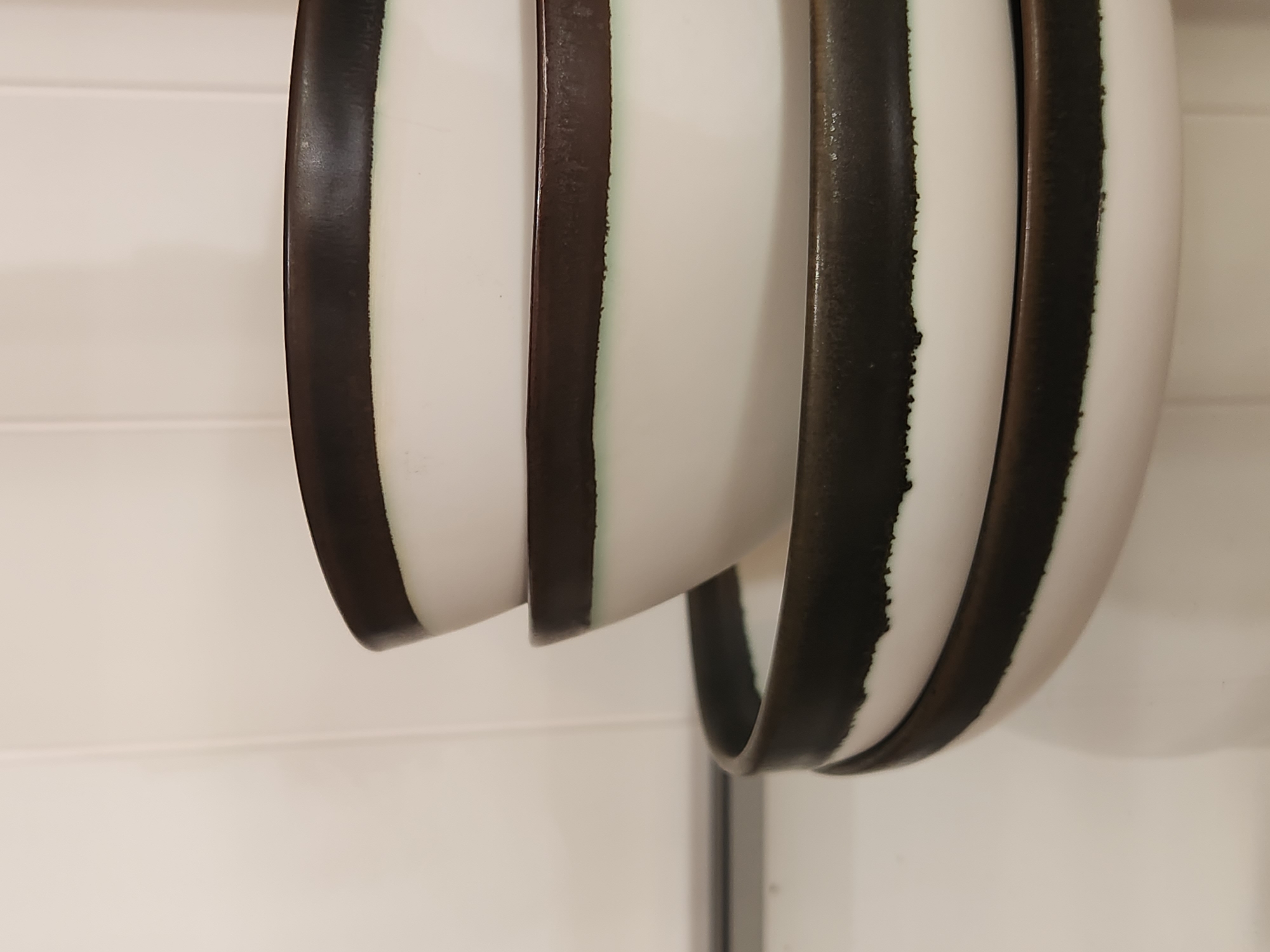 The media file shows a close-up, abstract view of multiple white ceramic bowls with thick, dark rims. The bowls are stacked or nested, creating layers of curved surfaces. The dark rims have a rough, textured appearance with irregular edges where the dark color meets the white ceramic. The lighting is neutral, suggesting an indoor setting. The background is a plain, light-colored wall with horizontal lines, possibly indicating paneling or wallpaper. There are no people visible, and the focus is solely on the texture and form of the ceramic bowls. The scene appears to be static, with no specific activity taking place. The location context is Maitland, United States of America. There is no visible text.