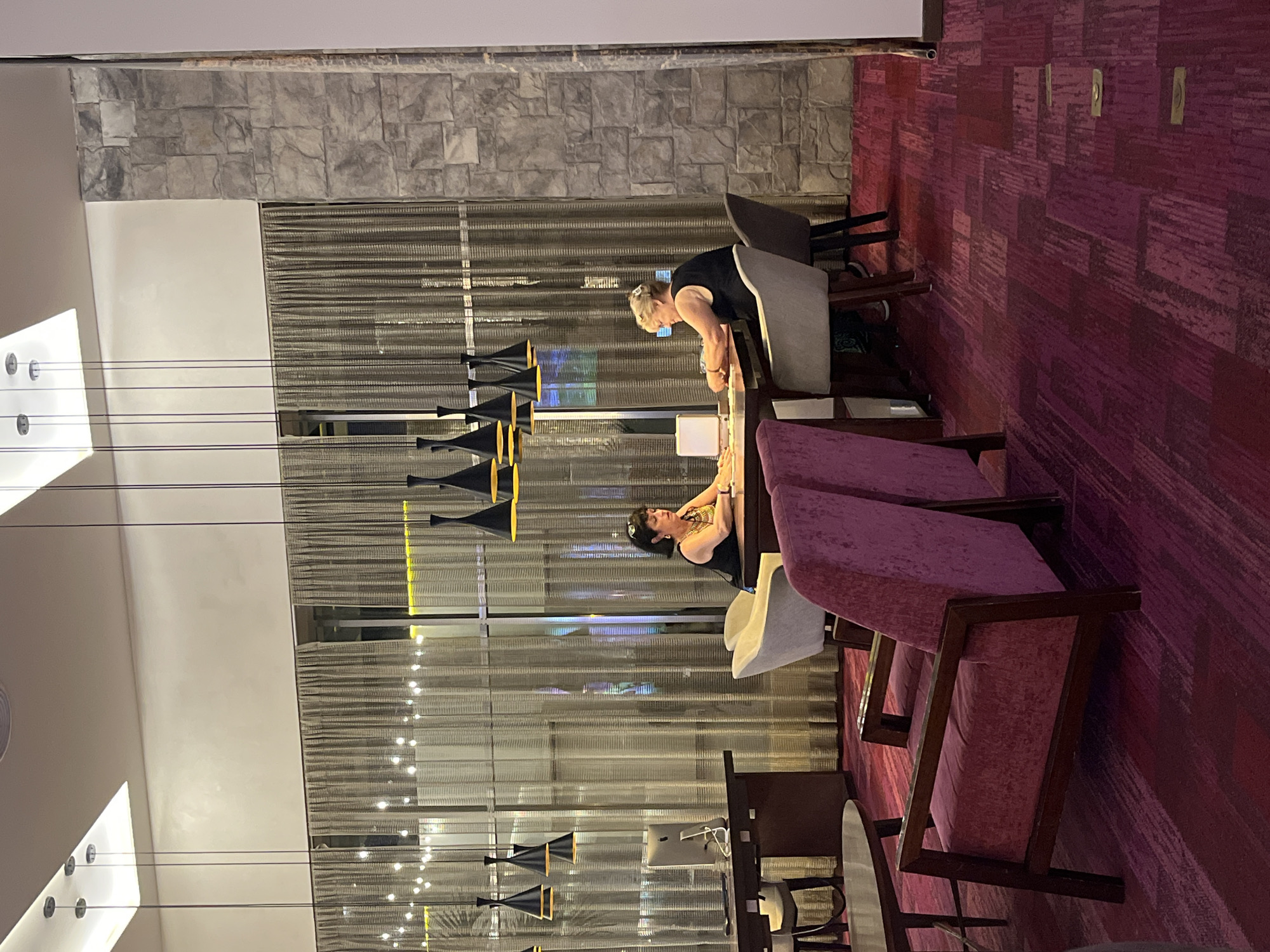 The interior of a public space in Playa del Carmen, Mexico, features two light-skinned women standing at a table, possibly a buffet, with one woman bent over the table. The women are wearing casual attire, with one in a black tank top and gold jewelry. The background includes a patterned wall, a stone wall, and a window covered by a thin curtain. In the foreground, there are chairs upholstered in pink fabric with dark wooden frames, and a contemporary chandelier hangs from the ceiling.