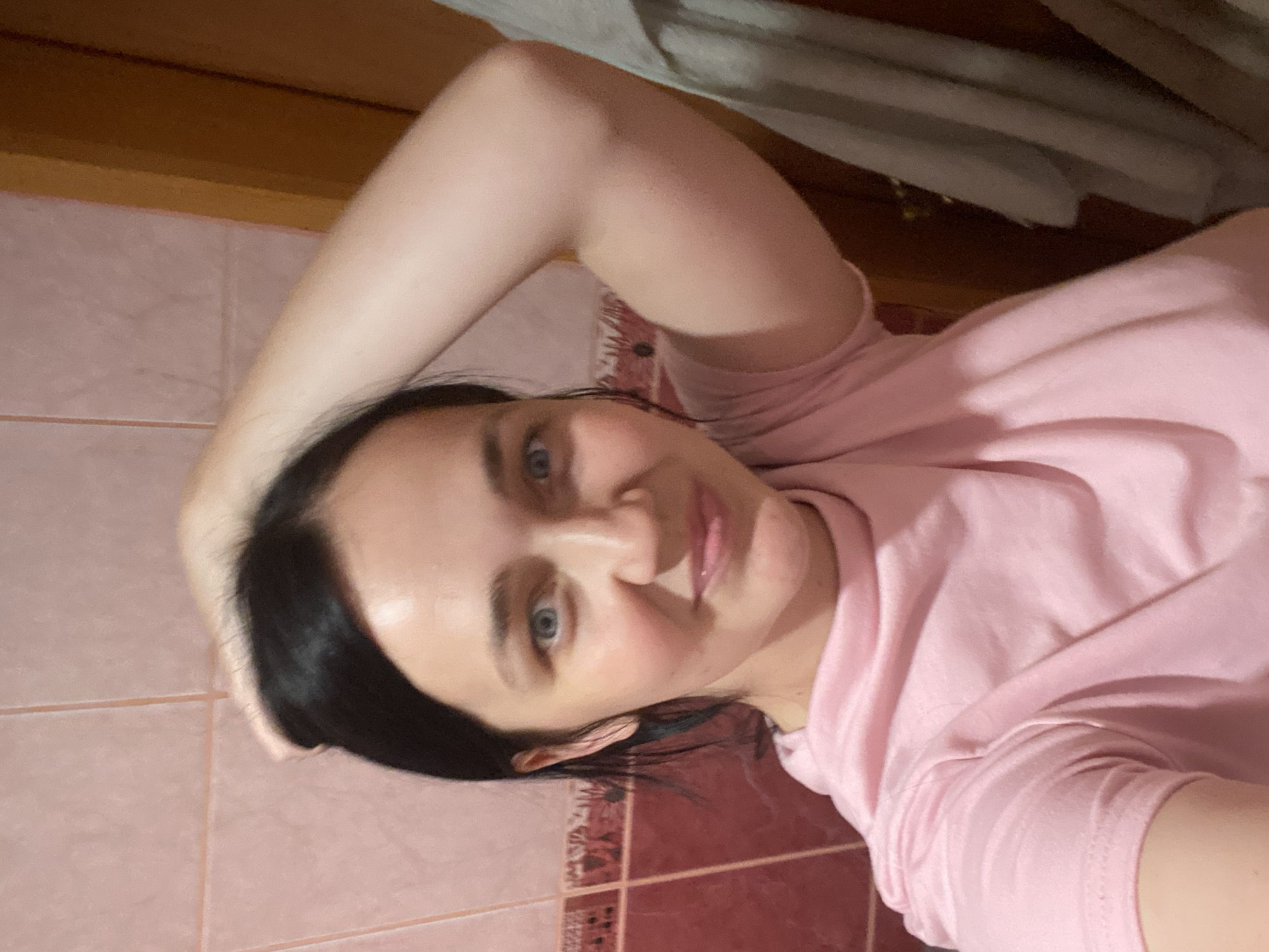 In a close-up, indoor shot with a shallow depth of field, a young woman with dark hair and blue eyes is shown from the chest up. She is wearing a pink t-shirt and has her right arm raised, with her hand resting on the back of her head. Her gaze is directed at the camera with a slight smile. The background consists of pink tiled walls with a decorative border, and a wooden doorframe to the right. A light-colored garment hangs on the right side of the frame. The lighting suggests an indoor environment, possibly a bathroom.