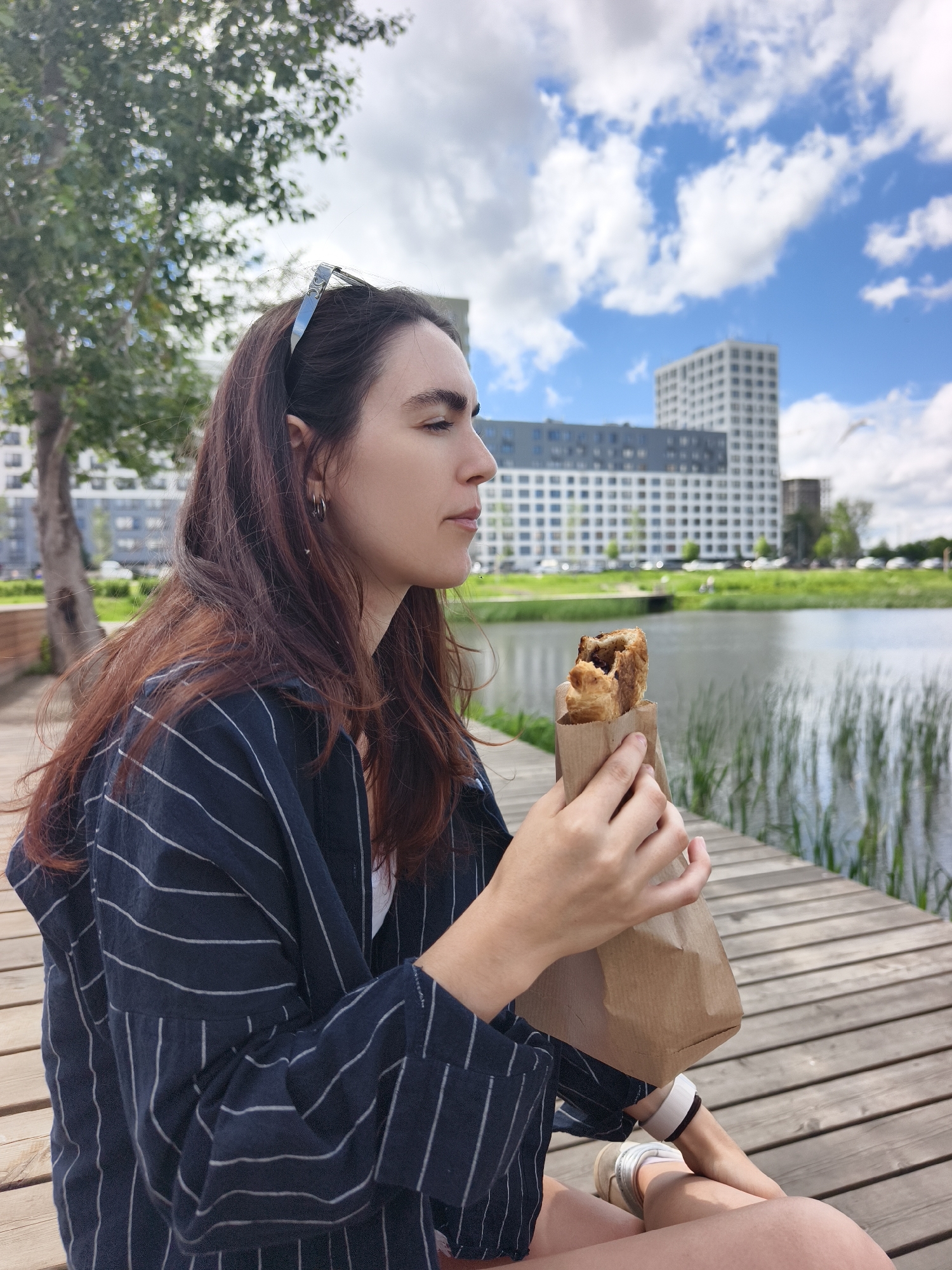 A young woman with long, dark brown hair is sitting on a wooden deck next to a body of water, holding a pastry wrapped in brown paper. She is wearing a dark blue and white striped shirt and white sneakers. In the background, there are modern buildings, trees, and a bright blue sky with white clouds. The location appears to be a park or waterfront area in the city of Tyumen, Russia.