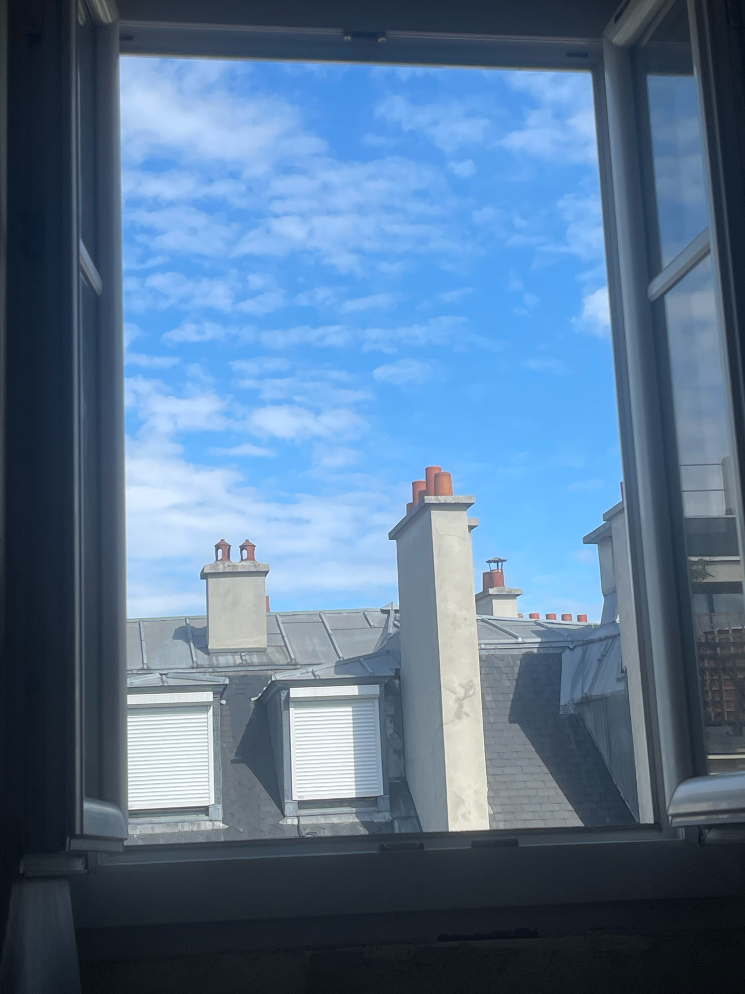 This is an eye-level, medium shot of a view through an open window in Paris, France. The foreground is dark, showing the bottom of the window frame and sill. The main subject of the image is a bright blue sky with scattered white clouds. Below the sky, the rooftops of Parisian buildings are visible, characterized by their grey slate tiles and various chimneys. The rooftops are mostly made of grey, mansard-style roofs with prominent chimneys. Some of the chimneys are topped with reddish-orange terracotta pots. In the middle ground, two windows with white roller blinds are visible on a building. The overall impression is a sunny day in a European city, seen from a residential window. There are no people or animals in the frame.