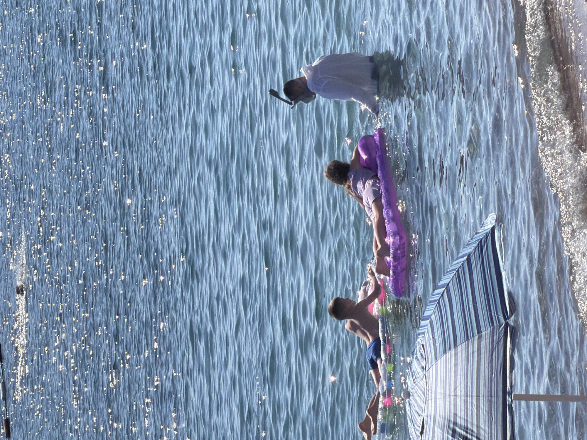 The image depicts a sunny day at a beach, likely in Petrovac centar, Montenegro, characterized by clear blue water with sparkling sunlight reflections on its surface. In the water, three individuals are visible. To the left, a young boy lies on his stomach on a colorful inflatable raft. In the center, another child, possibly a girl, is also on her stomach, resting on a purple inflatable raft. To the right, a person wearing a white shirt and a snorkeling mask stands in the water, facing the two children. In the foreground, a portion of a beach umbrella with blue and white stripes is visible, anchored in the shallow water. The overall scene conveys a relaxed summer atmosphere, with people enjoying the water and sun.
