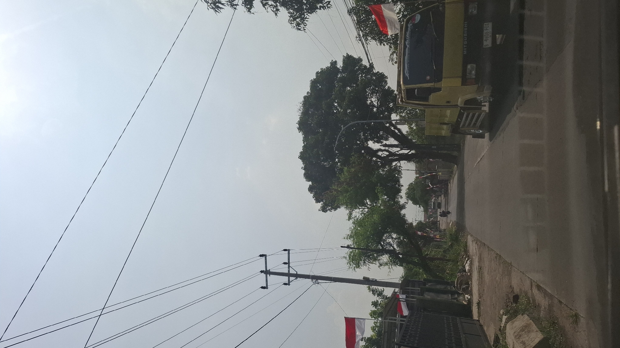 The image depicts a street scene in Sukasari, Indonesia, under a bright, overcast sky. Several utility lines crisscross the upper portion of the frame, supported by a utility pole on the left side of the road. Indonesian flags, red and white, are visible on poles along the side of the street. A yellow Mitsubishi truck is parked on the right side of the road, facing away from the camera. The road itself is paved and appears to be a residential street with houses and trees lining both sides. In the distance, a motorcycle is seen on the road. The overall impression is that of a typical Indonesian street scene on a clear day, possibly near a residential area.