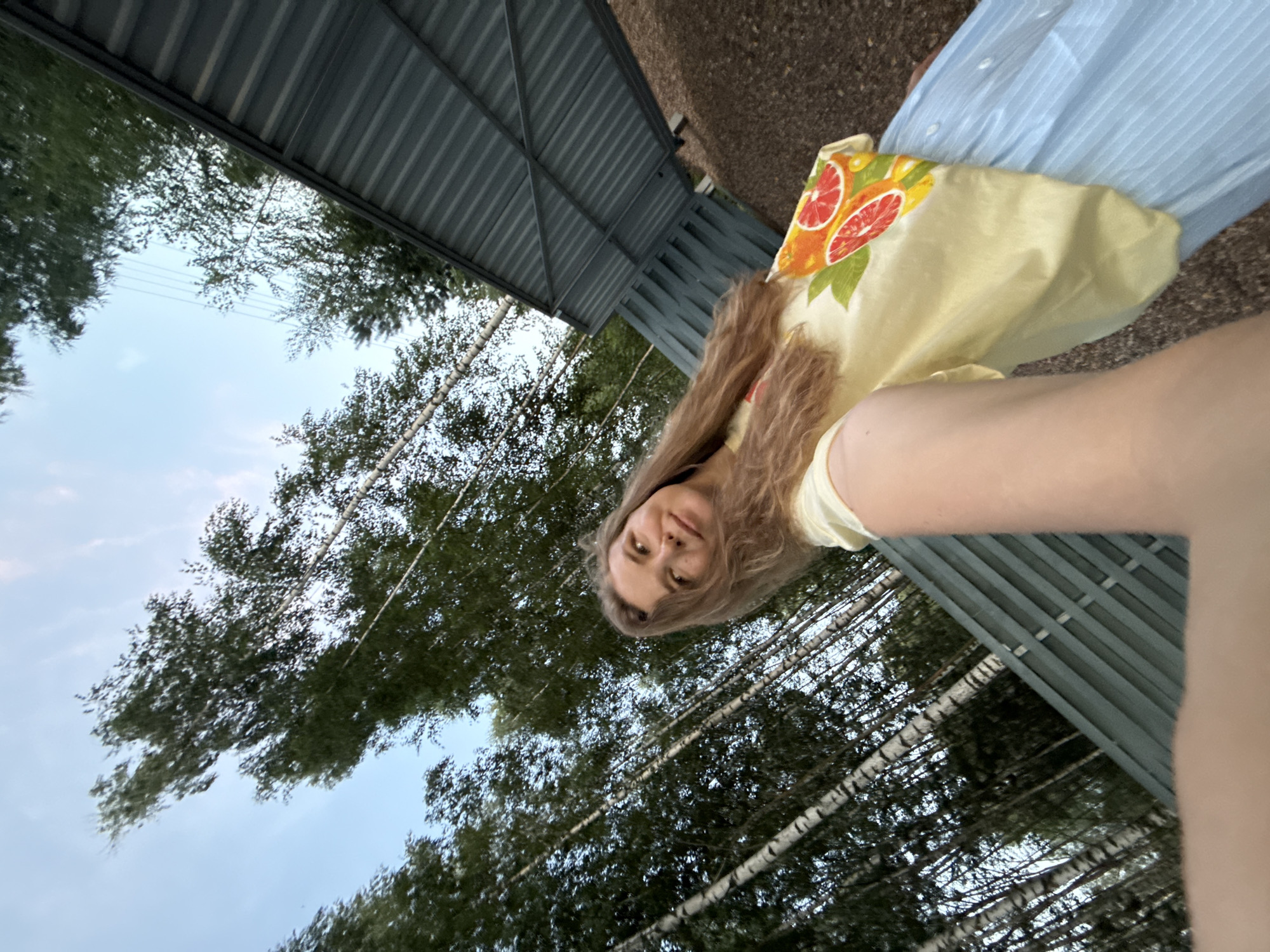 The image is a low-angle selfie taken outdoors. A young woman with long, wavy blonde hair is looking up and to her right. She is wearing a yellow t-shirt with a graphic print of sliced citrus fruits and a light blue pleated skirt. The background is a grove of birch trees with their distinctive white and black bark, and a portion of a gray corrugated metal structure is visible on the right side of the frame. The sky is partly cloudy. The location is the city of Prokudino, Russia.