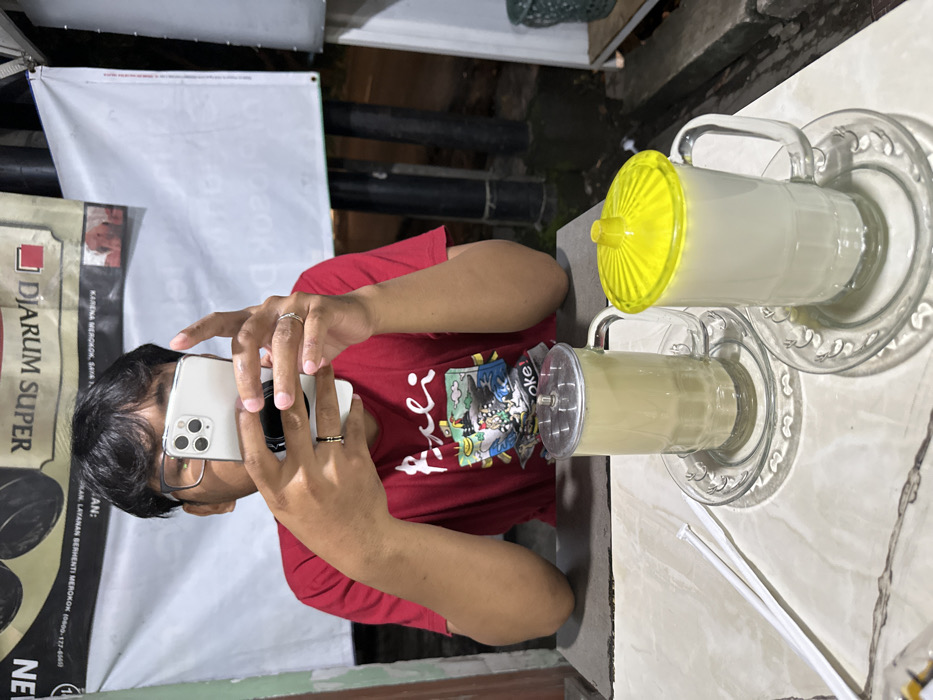 The image captures a person taking a photograph of two drinks on a table, presented in a sideways orientation. The main subject is a person, likely male, with short dark hair and glasses, wearing a red short-sleeved t-shirt with a colorful graphic design on the chest that includes the partially visible text "oke!". He is holding a white smartphone, which appears to be an iPhone with three camera lenses, in both hands, positioned in front of his face as if taking a picture or video. A black pop-socket or phone grip is visible on the back of the phone. He wears a thin silver-colored ring on his right ring finger and a black and gold ring on his left ring finger.
On the table in front of him are two clear glass mugs, each filled with a light beige or off-white beverage. The mugs are placed on matching clear glass saucers with ornate, ridged edges. The mug closer to the viewer has a silver-colored, flat lid with a small central opening, while the mug further away has a bright yellow, ribbed, dome-shaped lid. White plastic straws are visible lying on the light-colored, possibly marble or tiled, table surface next to the mugs.
In the background, to the left of the person, a white banner or sign is visible, partially cropped. Prominent text on this banner includes "DJARUM SUPER," a brand of cigarettes, followed by smaller text in Indonesian: "KARENA MEROKOK, SAYA" (Because smoking, I...) and "LAYANAN BERHENTI MEROKOK 0800-177-6500U" (SMOKING CESSATION SERVICE 0800-177-6500U). Other partial texts visible are "AN:", "NE," and the number "8." The setting appears to be indoors or a covered outdoor area, possibly a cafe or restaurant, with relatively dim lighting suggesting it might be evening.